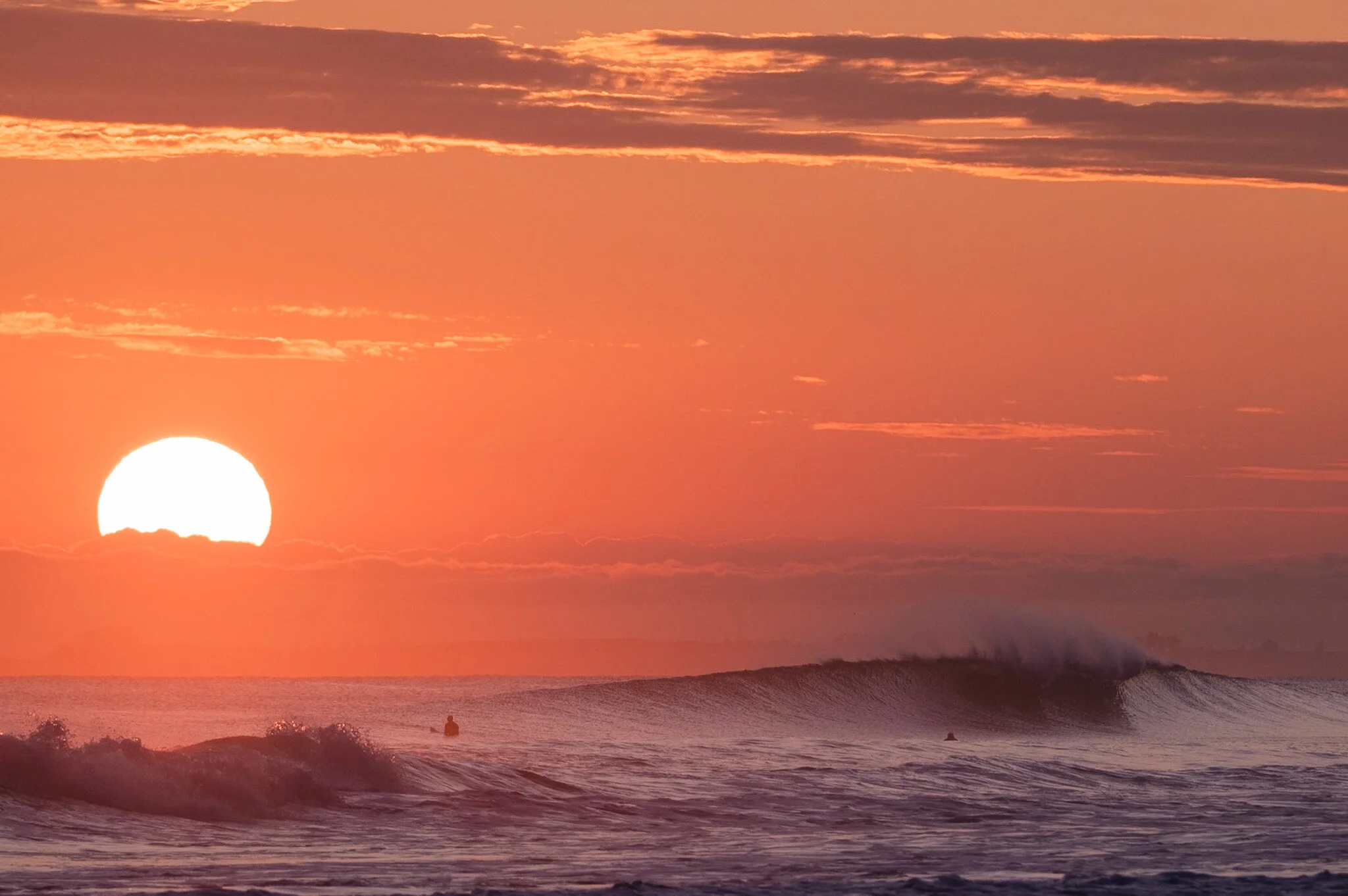 A massive wave crashes onto the shore of Mount Maunganui beach during Cyclone Tino, while the sun rises in the background, creating a stunning contrast between the fiery sky and the frothy sea. This beach landscape photo was taken by ocean photograph