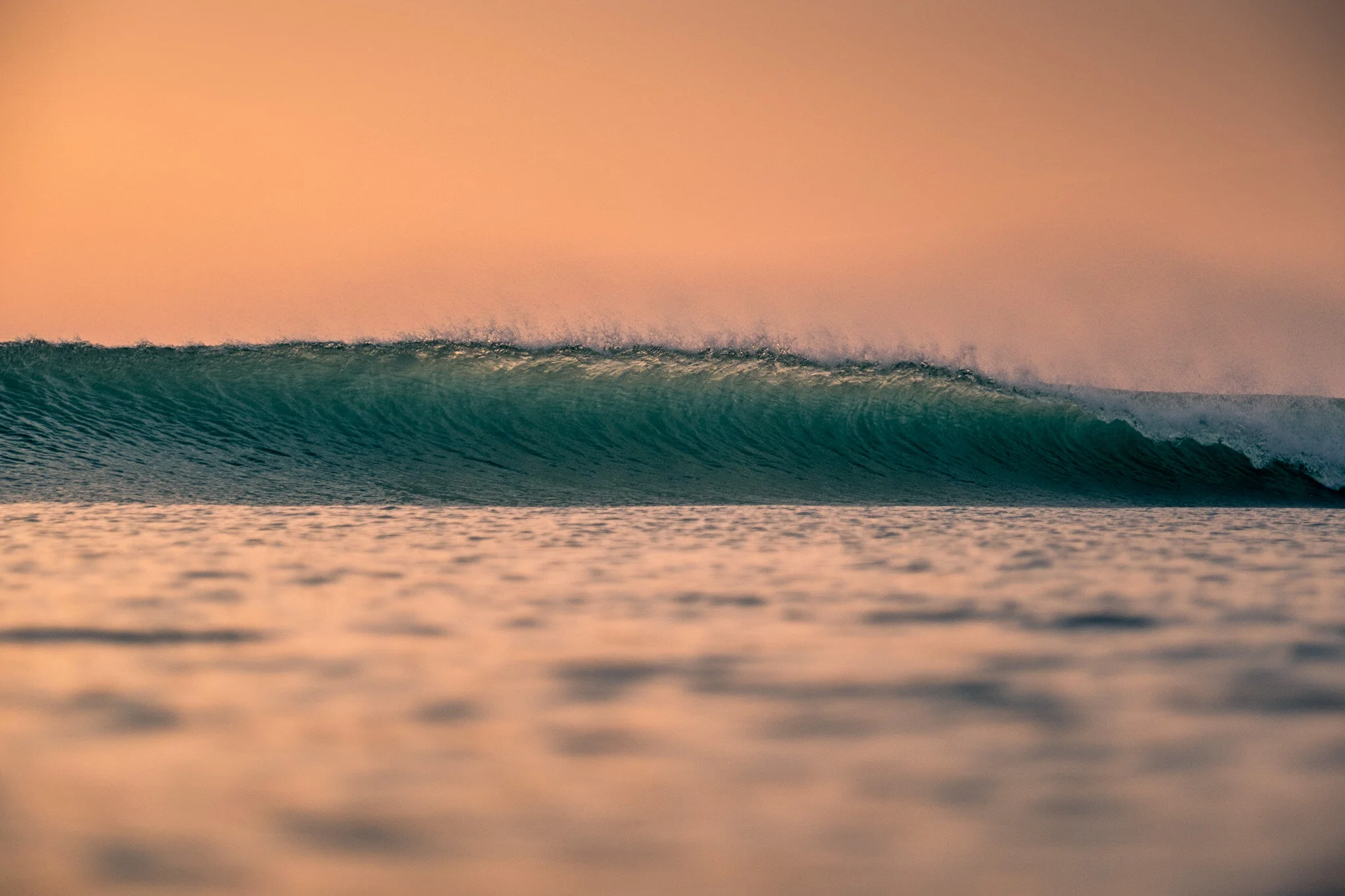 A perfect wave at sunrise captured in a photo, with a peach-colored sky in the background and a turquoise wave in focus in the foreground. A breathtaking and serene moment in time showcasing New Zealand surf by Lou Lou B Photo.