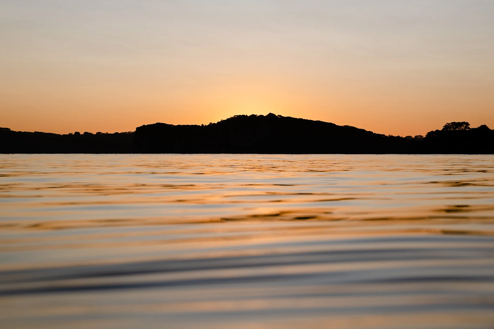 Sunrise over Whitianga Beach, New Zealand. Shakespeares cliff is silhouetted in the background. The orange tones of the sky are reflected on the surface of the ocean in the foreground, taken at water level. A peaceful and serene beach landscape captu