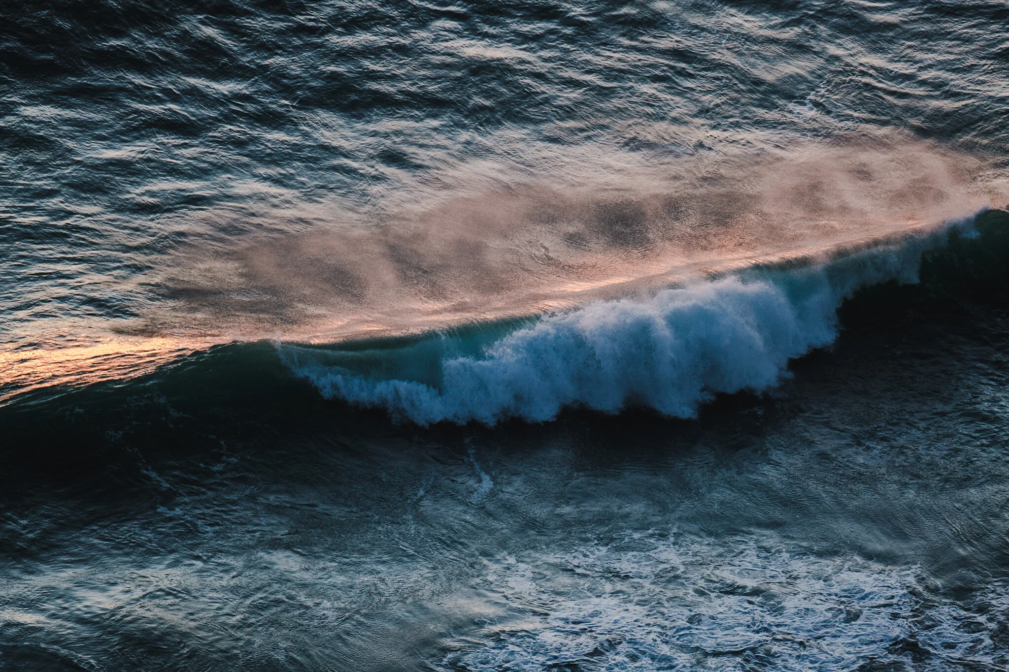 Photo of a wave captured from above, the ocean is a deep turquoise blue with a soft orange reflection on the whitewash. The shot is minimalist and focused on the interplay of colour and light. Photographed in Mount Maunganui by Lou Lou B Photo.