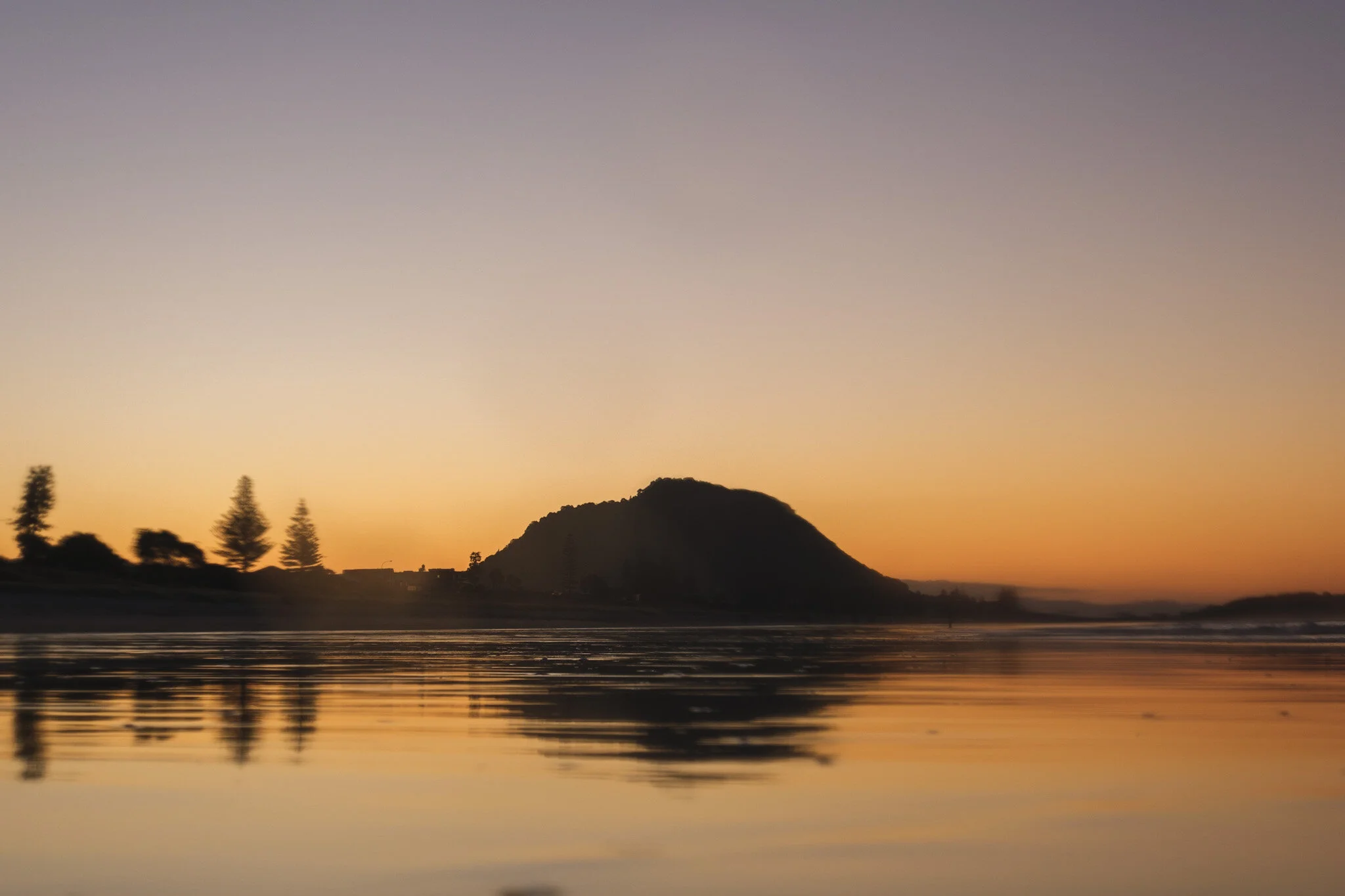 Silhouetted Mount Maunganui at sunset, casting golden reflections on the low tide beach, with an orange sky as the backdrop. A peaceful beach landscape captured by ocean photographer Lou Lou B Photo.