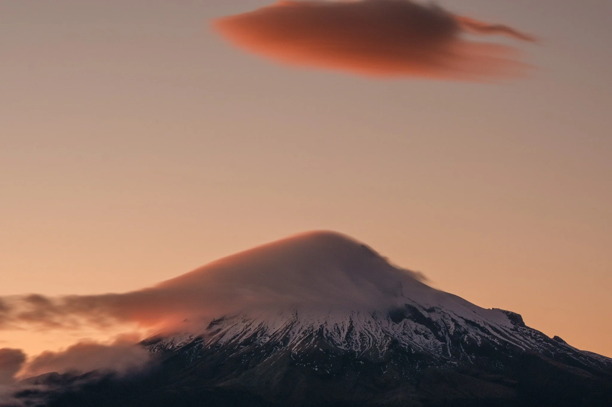 A stunning photograph of Mount Taranaki captured by New Zealand landscape photographer Lou Lou B Photo. The mountain stands tall and proud in the center of the frame, with its snowy peak visible against the soft pink hues of the sky. The use of a slo