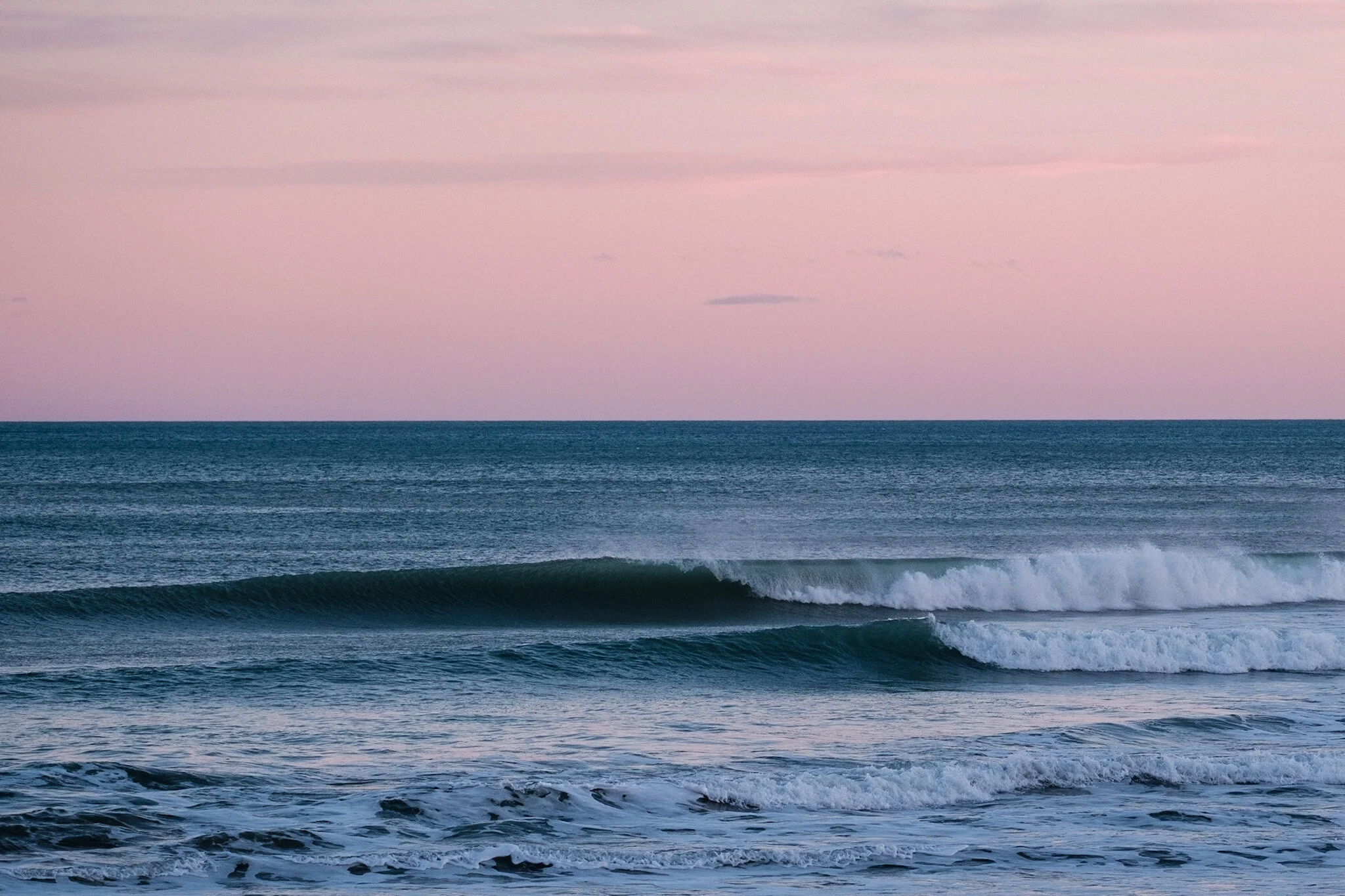 Two gentle waves roll onto the shore at Makarori Beach in Gisborne as the sun sets. The eastern sky is painted with pastel pinks and purples while the ocean appears as a rich blue. Serene beach landscape photo by Lou Lou B Photo.