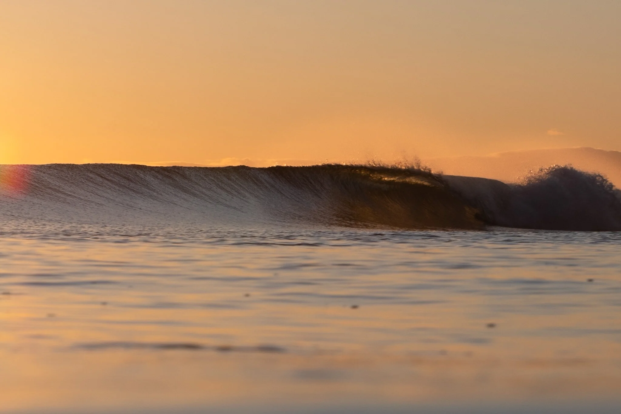 Sunrise at Mount Maunganui, a perfect wave breaks in the distance, its curl glowing with warm orange light. In the foreground, a rainbow-colored orb illuminates the scene, adding a magical touch to the serene atmosphere.