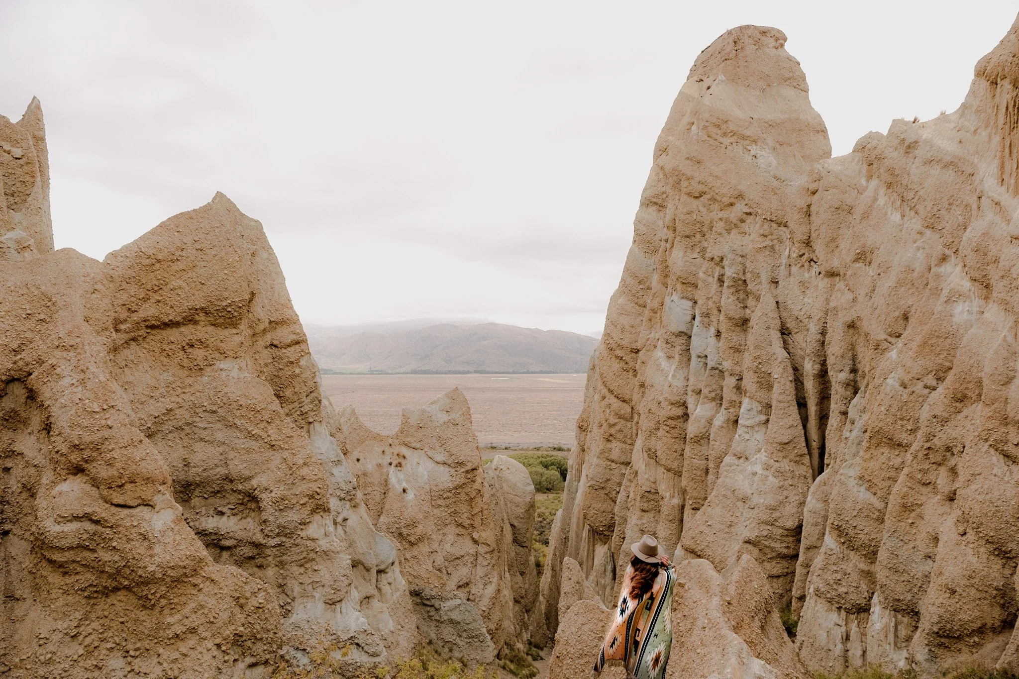 A person standing on the edge of Omarama Clay Cliffs in New Zealand, wearing an Aztec blanket and fedora hat. The person is looking out at the stunning natural landscape, with the rugged cliffs and rolling hills stretching out before them. The warm c