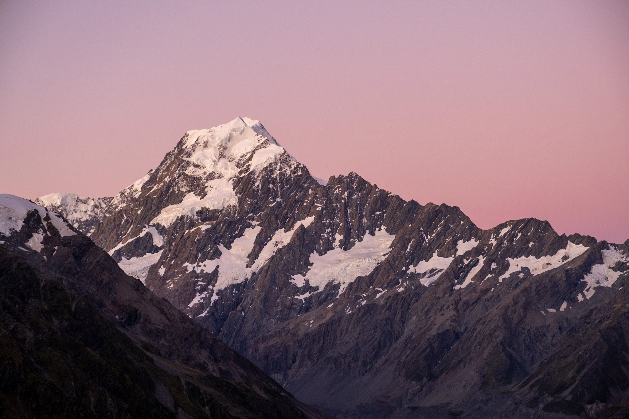 A stunning blue hour photograph by Lou Lou B Photo showcasing the majestic Mount Cook in New Zealand's National Park. The snow-capped peak of the mountain dominates the frame, with the pink hues of the after-sunset sky serving as a beautiful backdrop