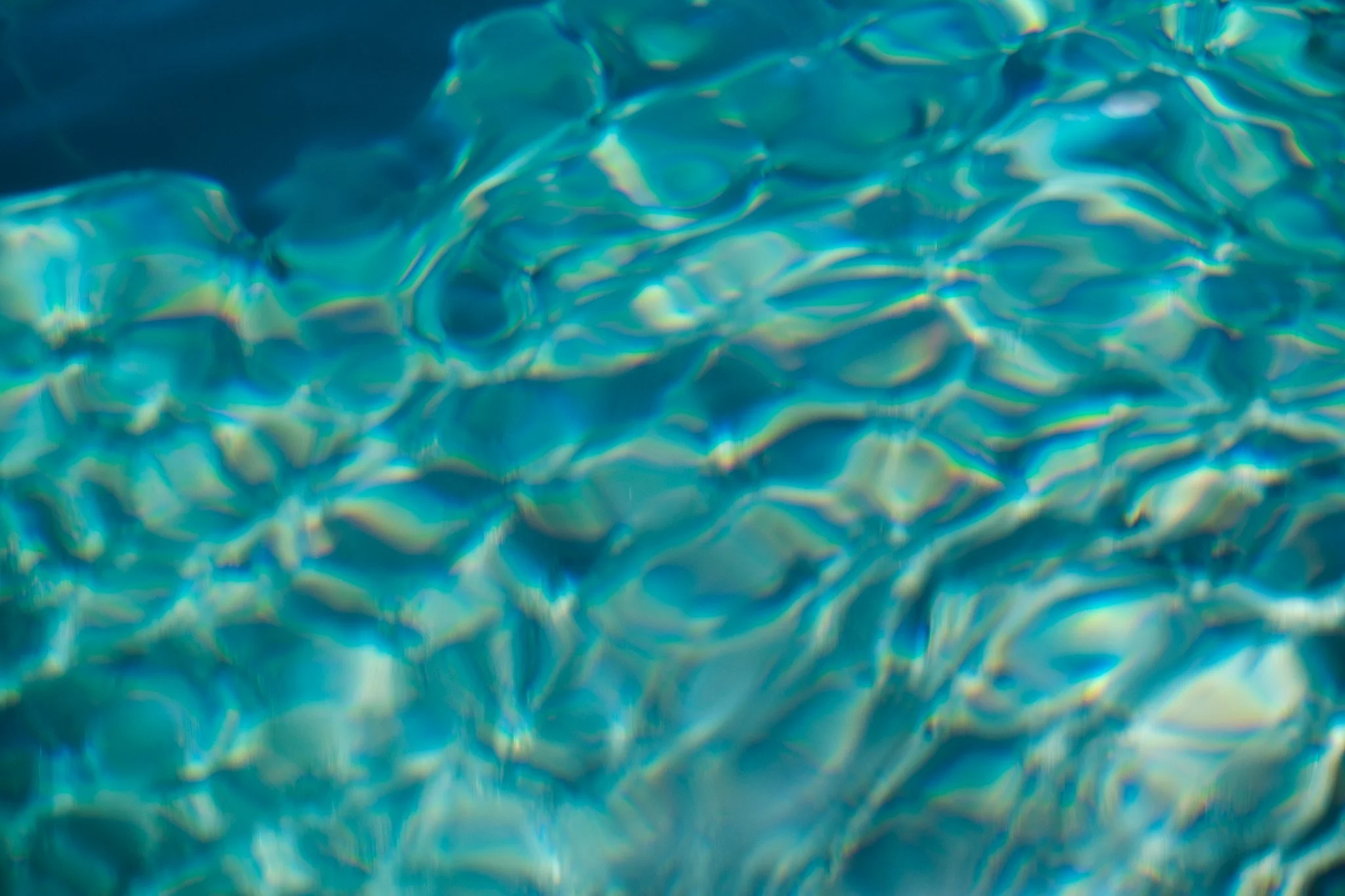 An aerial photo of the stunningly beautiful Blue Pools in Wanaka, New Zealand. The crystal clear turquoise water is the main focus, with soft ripples and reflections visible through the surface. The photo is shot from above, creating a dreamy, ethere