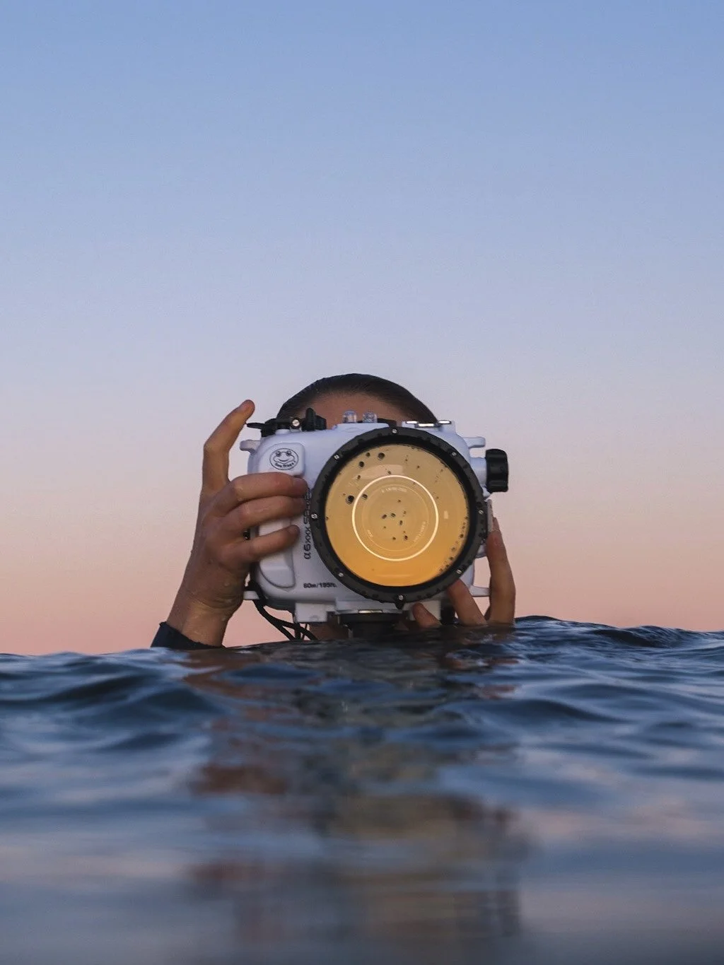 A photo taken by Lou Lou B Photo of a female surf photographer in the ocean in New Zealand, with a pastel sky behind her and the warm orange tones of sunset reflected in the lens port of her water housing.