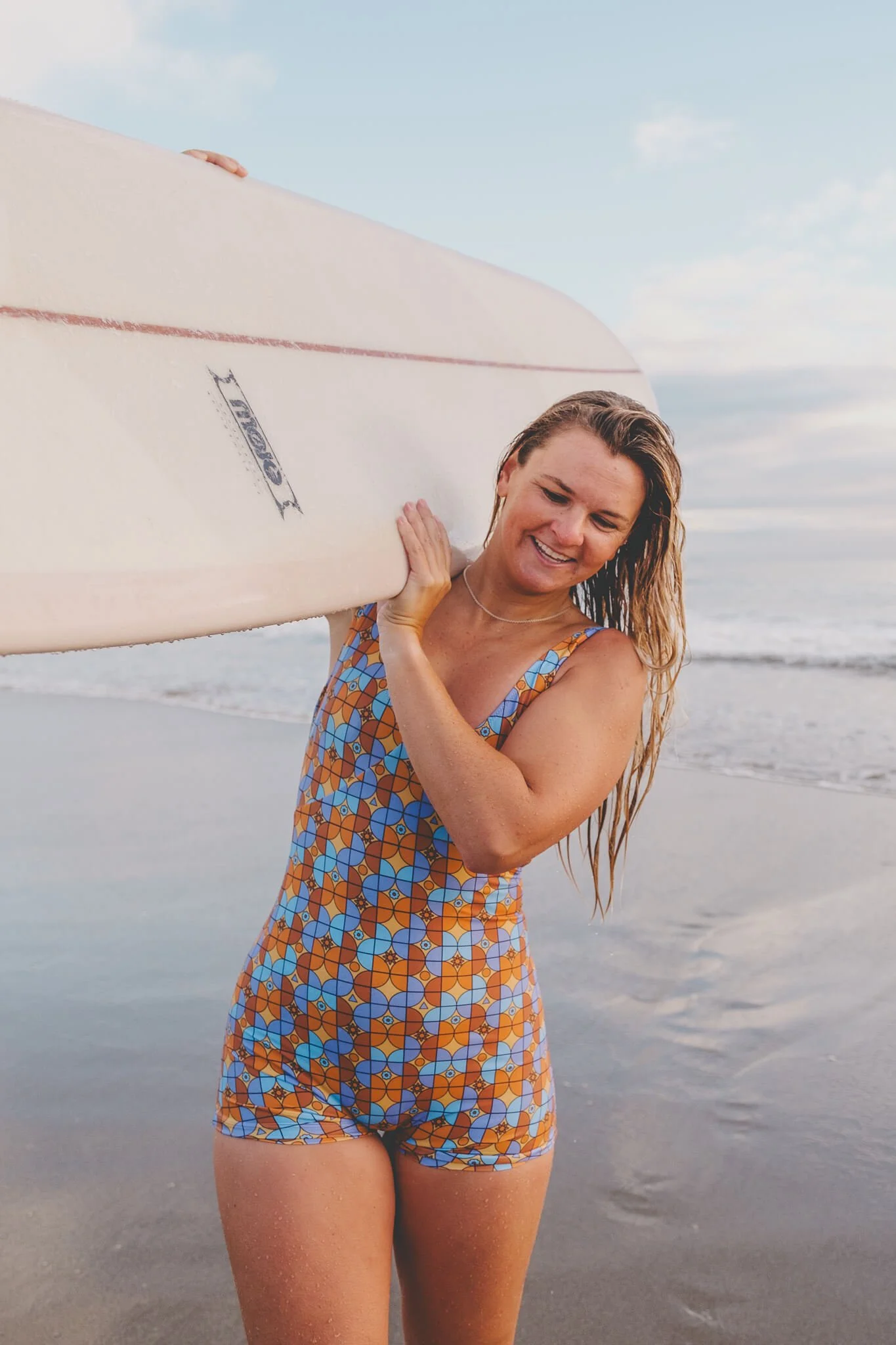  Smiling woman in a retro-patterned swimsuit carrying a surfboard along the shoreline after an early morning surf in Mount Maunganui. 