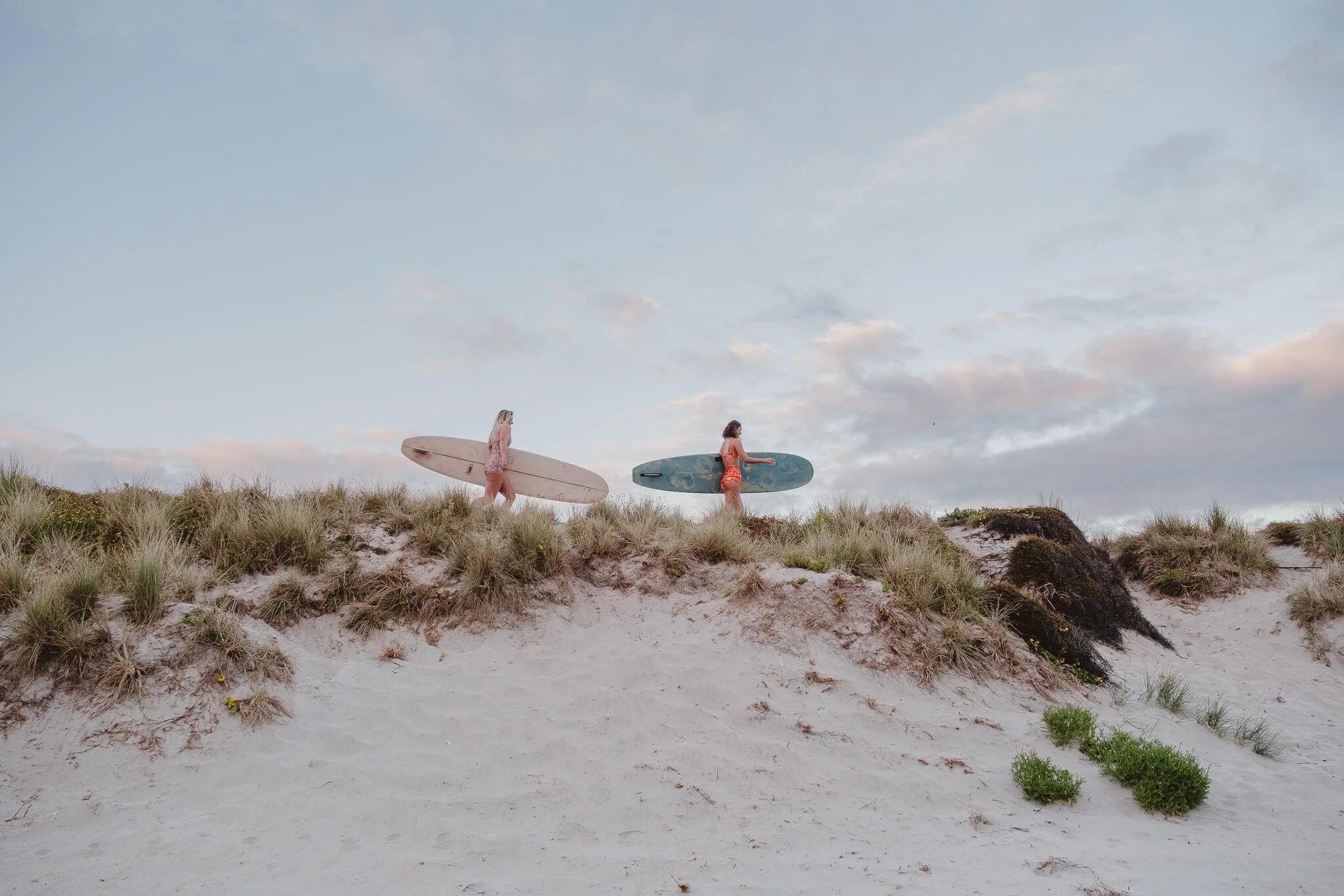 Two girls with surfboards walking across sand dunes