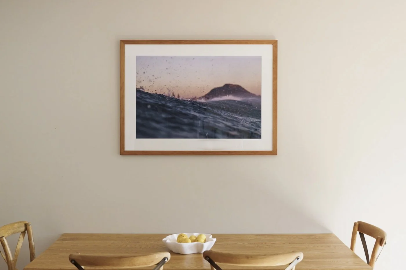 Framed fine art photograph of Mauao, Mount Maunganui, rising behind ocean waves at sunset, displayed above a wooden dining table.