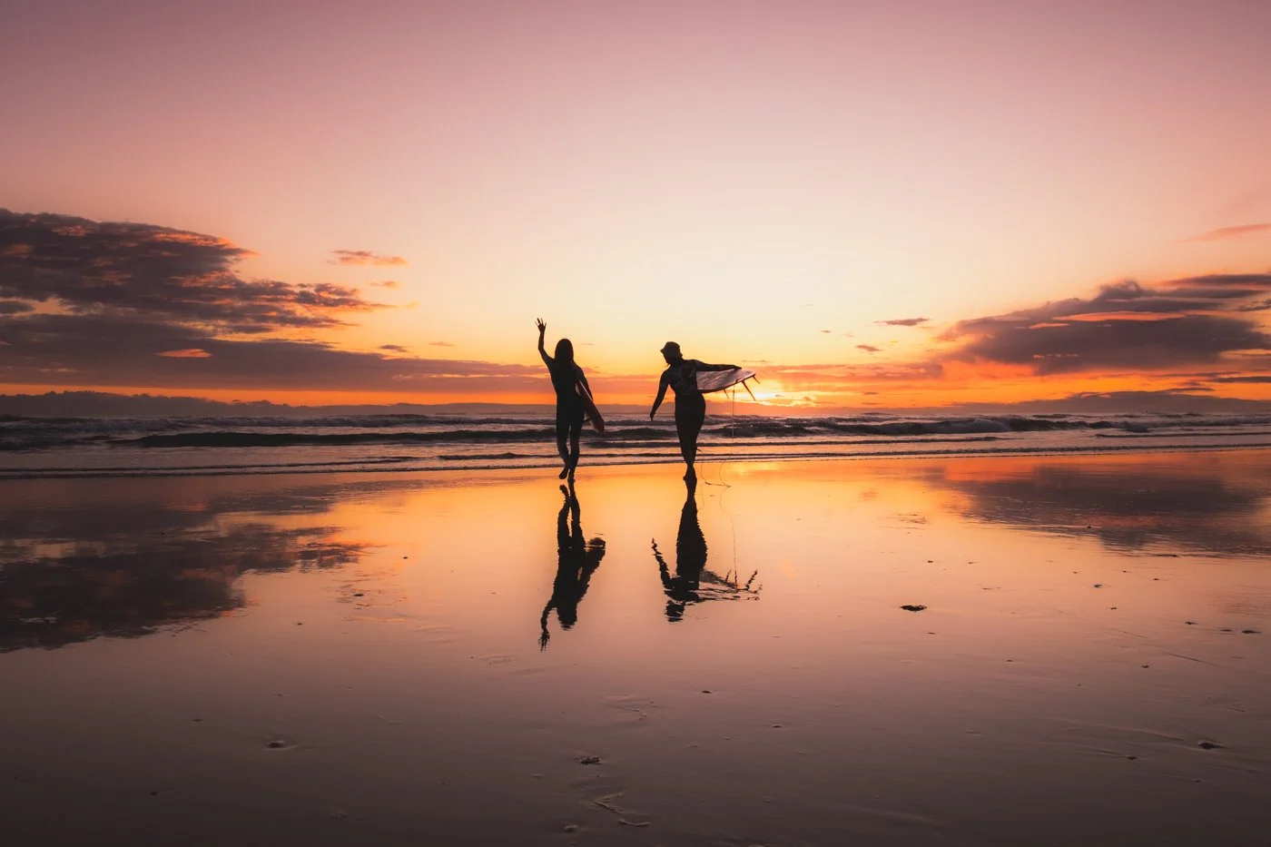 Surfers walking along reflective wet sand at sunrise in Mount Maunganui with pastel sky and ocean horizon