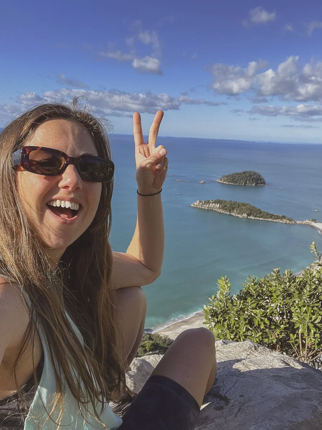 Smiling woman wearing sunglasses at the summit of Mauao, with a peace sign and views of the ocean and offshore islands.
