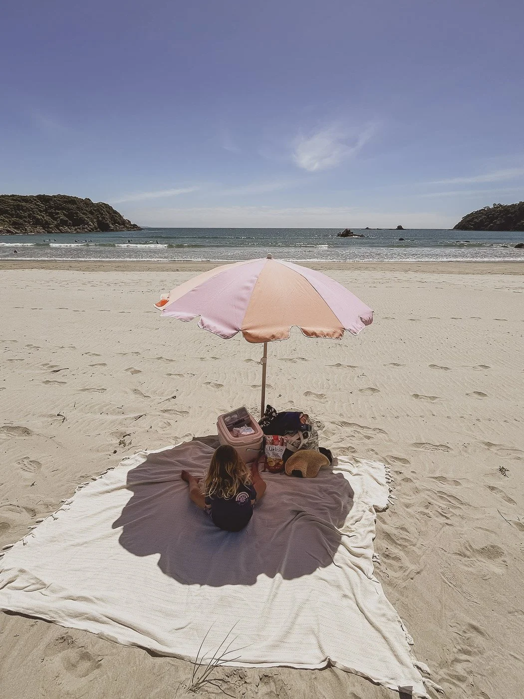 Pink beach umbrella shading a picnic blanket on white sand, with the ocean and rocky headlands in the background.