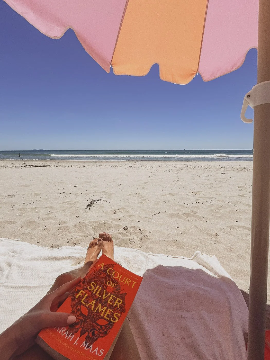 Relaxing on a beach towel beneath a pink umbrella, reading a book with a view of the sandy beach and ocean.