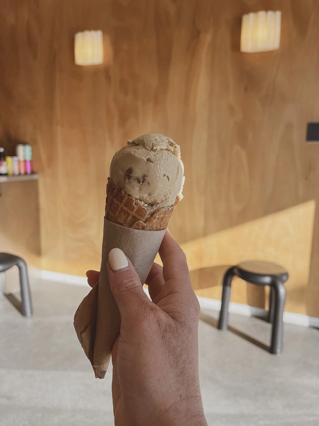 Hand holding a scoop of chocolate-coloured ice cream in a waffle cone inside a Mount Maunganui ice cream shop.