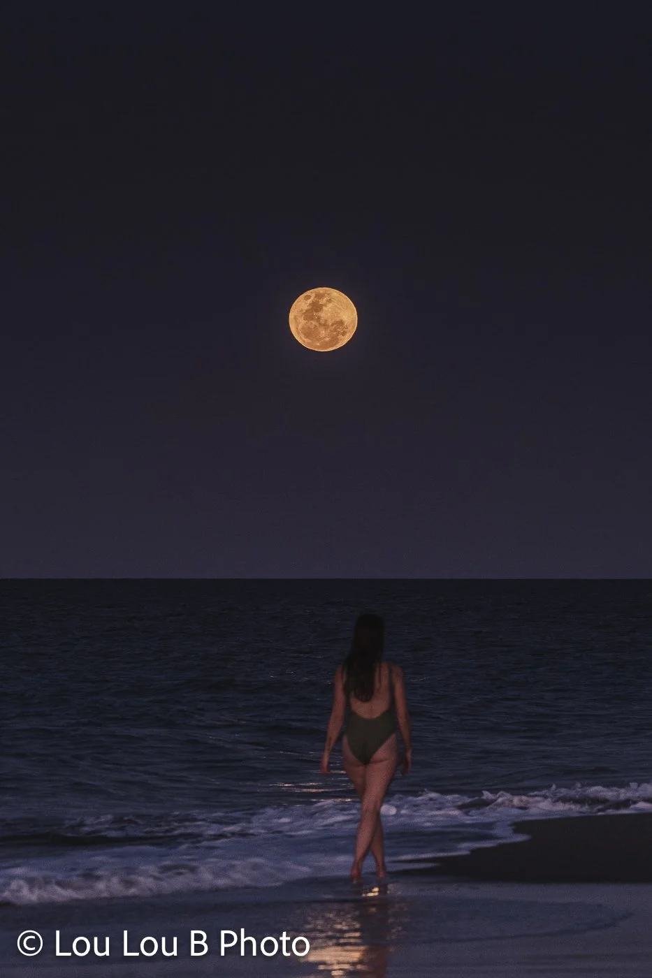 Woman walking along the shoreline beneath a small golden full moon at dusk.