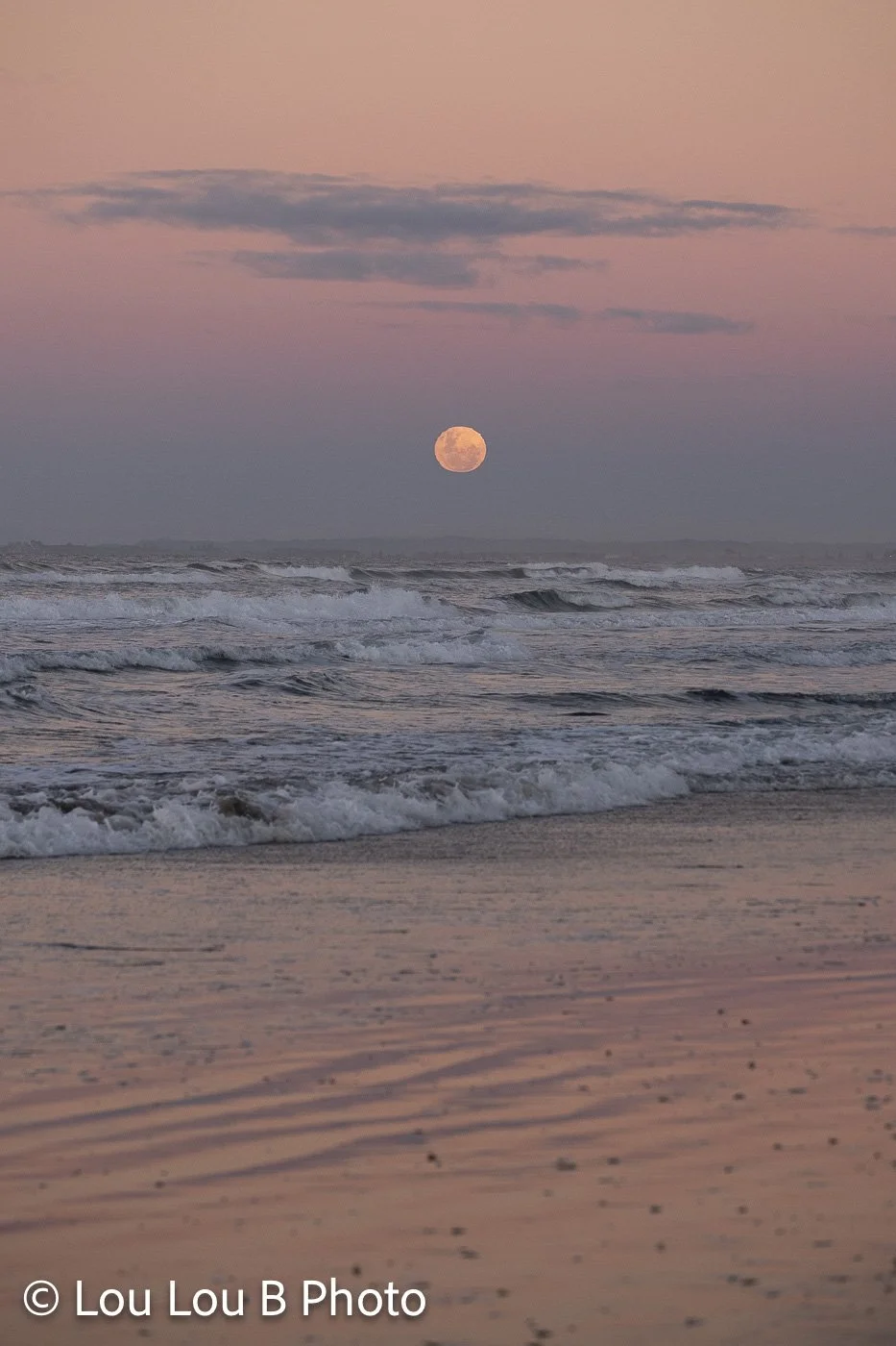 Small golden moon rising above breaking ocean waves and a sandy beach at dusk.