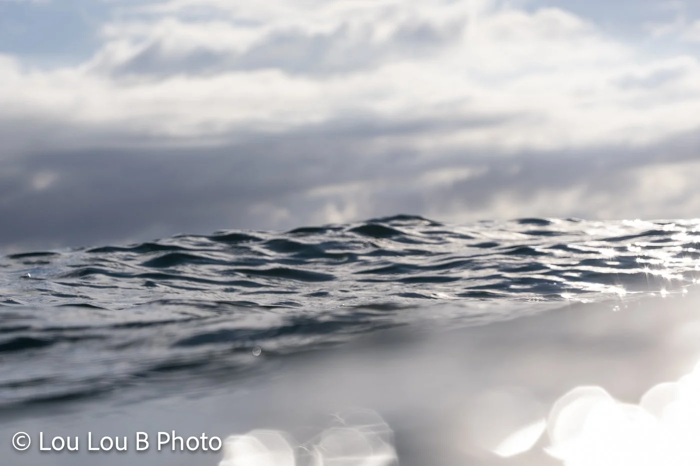 Low water-level view of blue-grey ocean swells beneath a cloudy sky with sunlight reflecting on the sea.