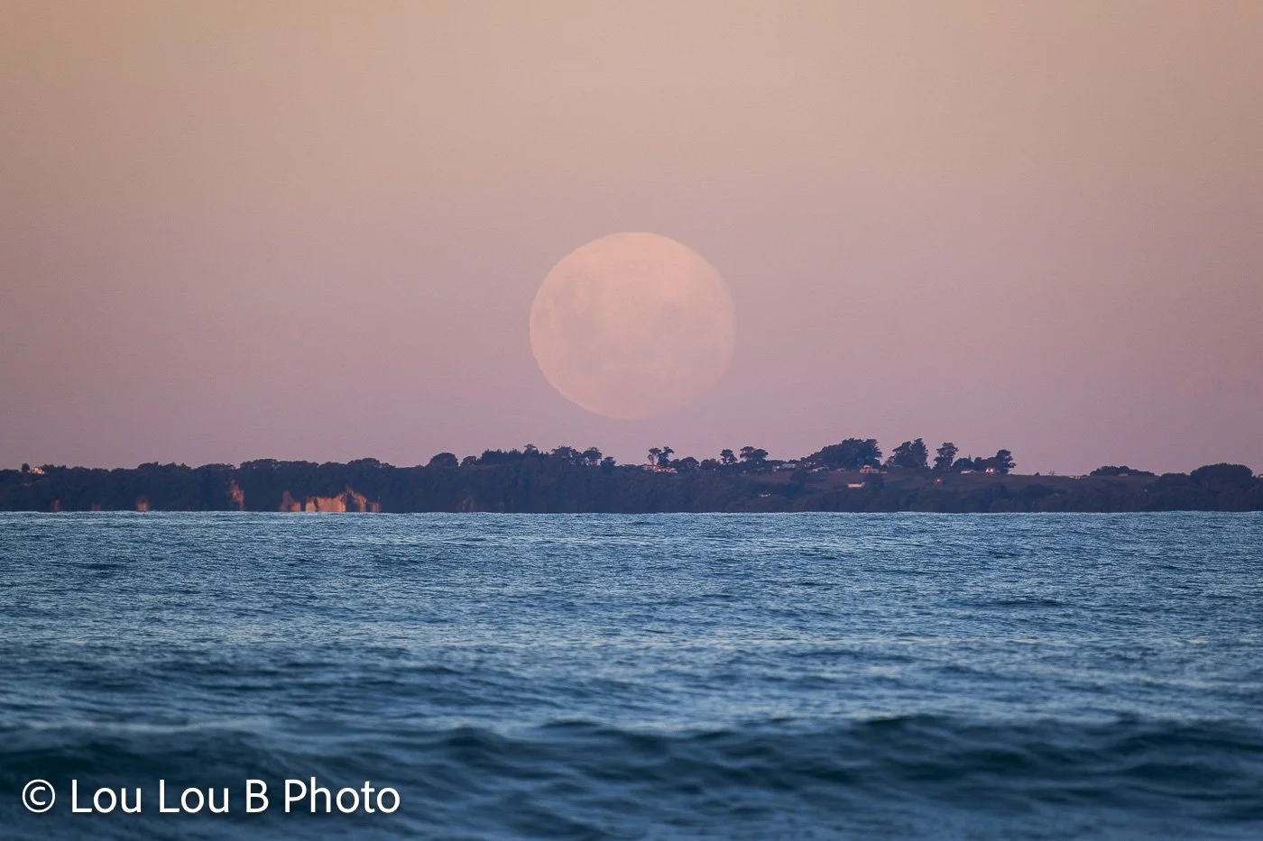 Pastel full moon rising above a distant island coastline and blue ocean at dusk.