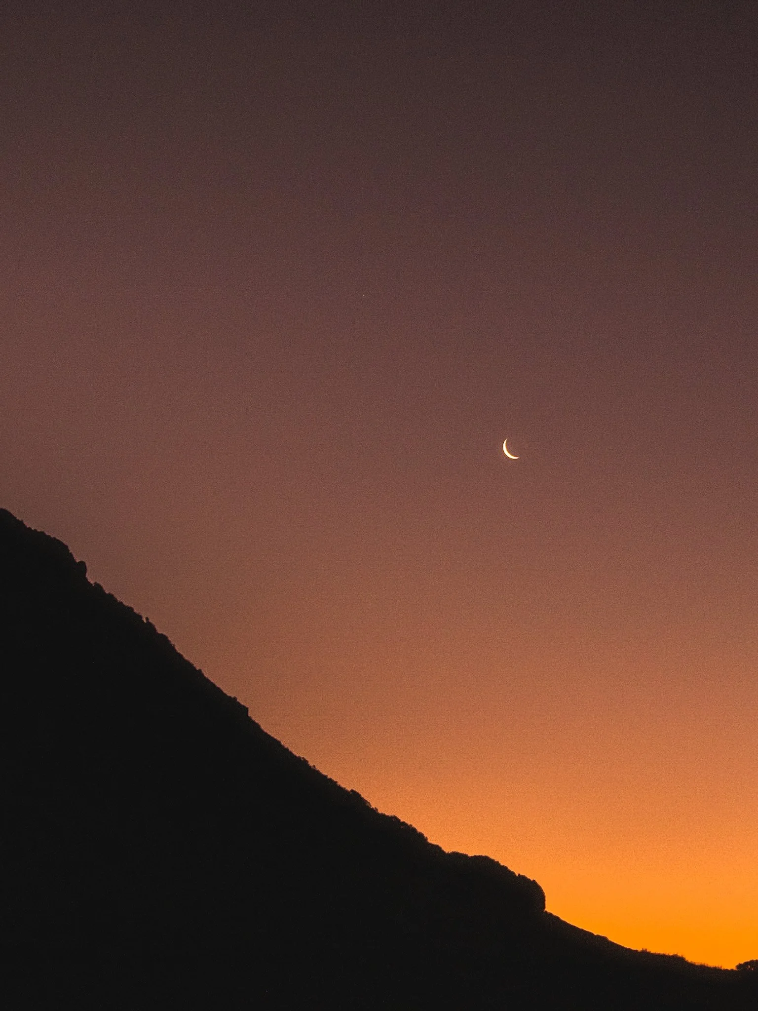 Immerse yourself in the captivating beauty of nature with this stunning photograph of a crescent moon shining against an autumnal orange sky, with the silhouette of Mount Maunganui in the left part of the composition. The warm and vibrant colors of t
