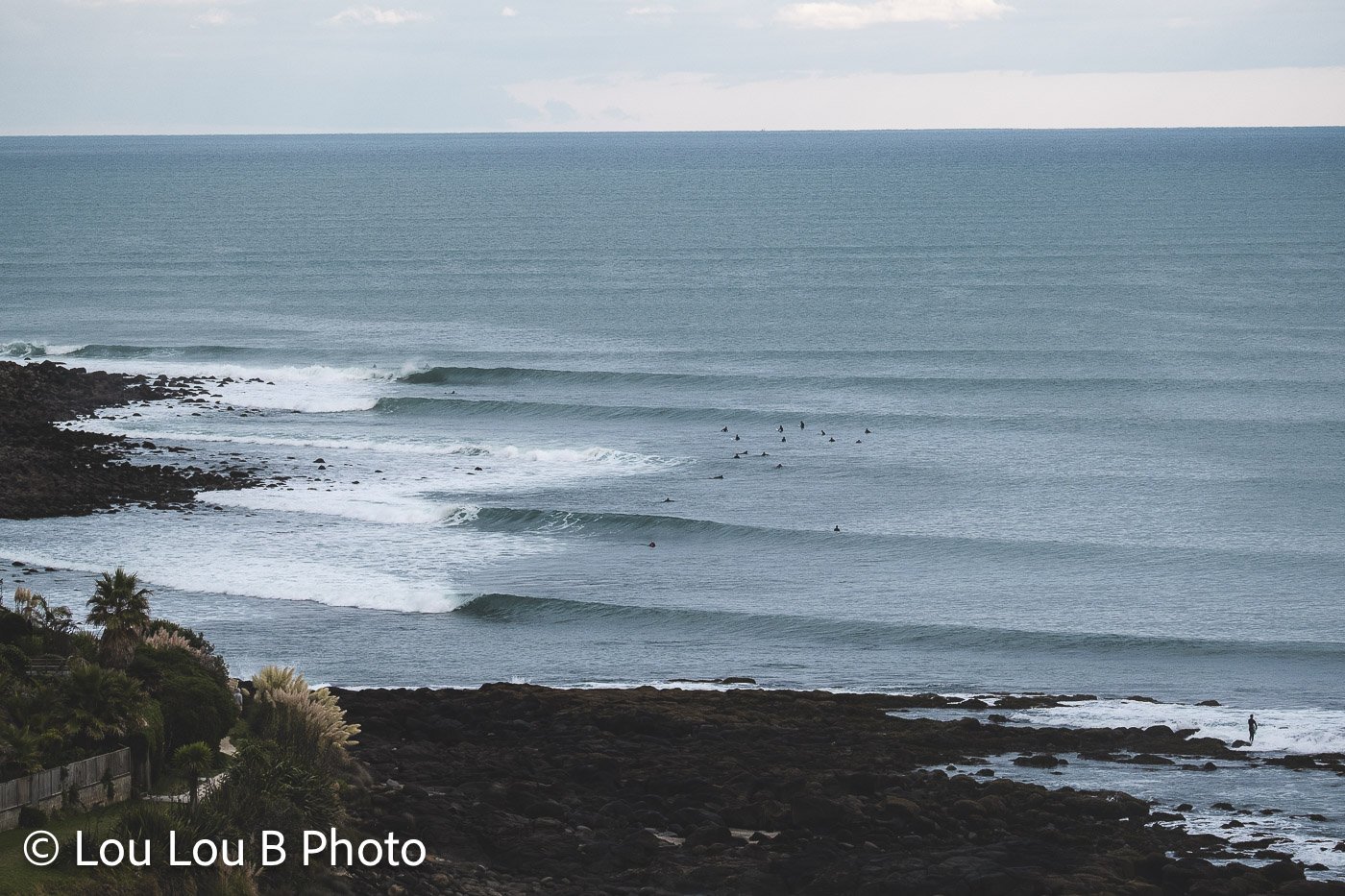 Elevated view of long ocean swell lines breaking along a rocky coastline with surfers in the water.
