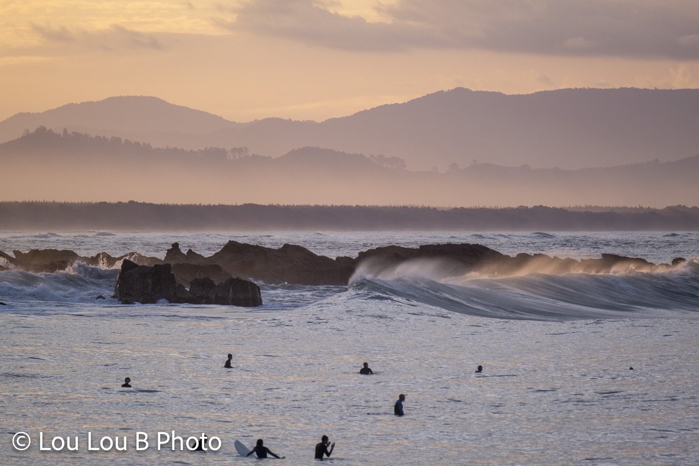 Surfers in the ocean at Mount Maunganui with waves and coastal hills in golden evening light