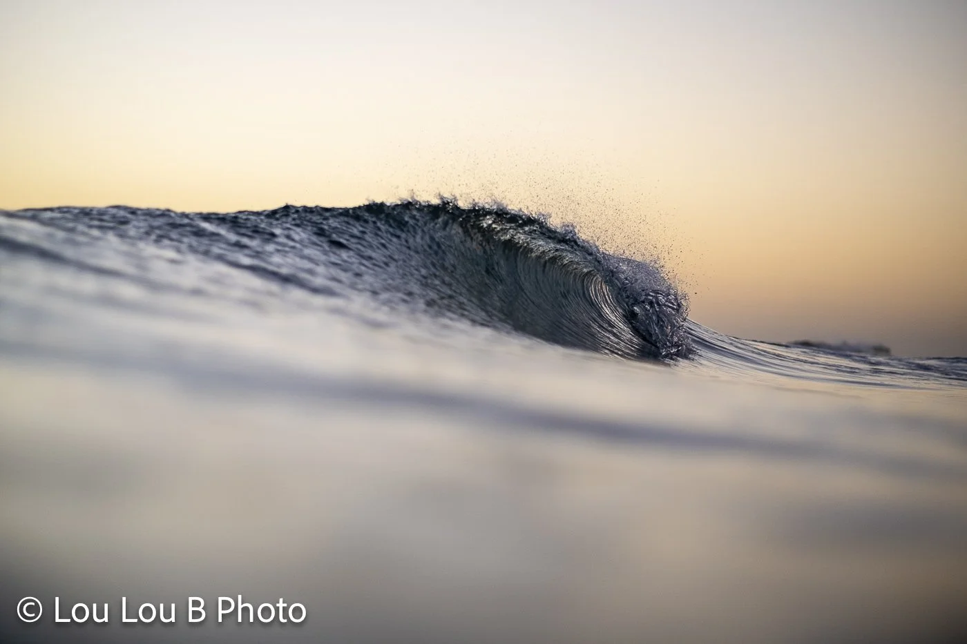 Close-up water-level view of a curling blue wave beneath a warm cream and peach sky.