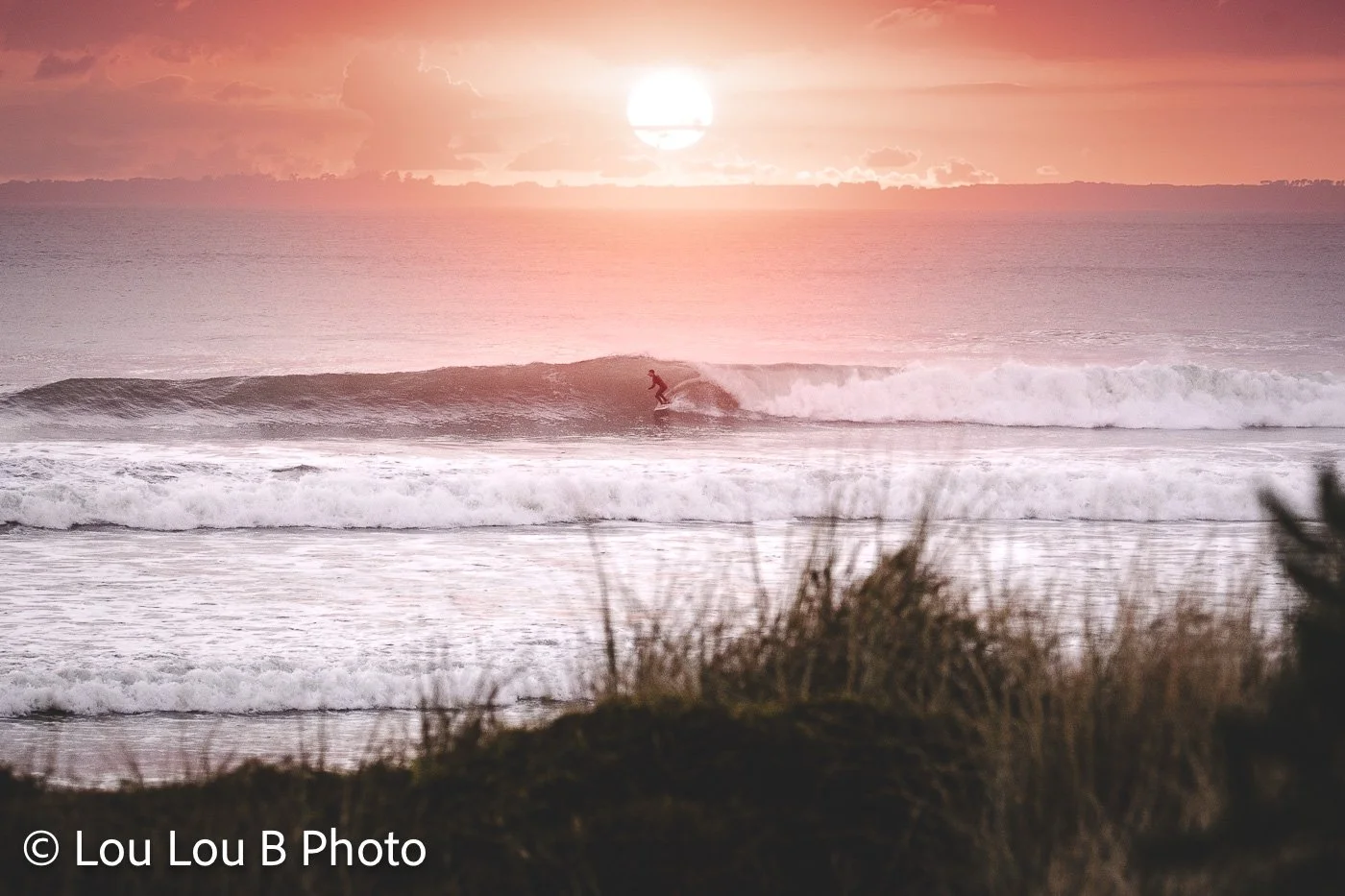 Breaking ocean waves beneath a glowing pink-orange sunset over the coast.