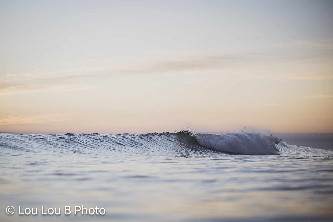 Small blue wave breaking across calm water beneath a soft peach and blue pastel sky.