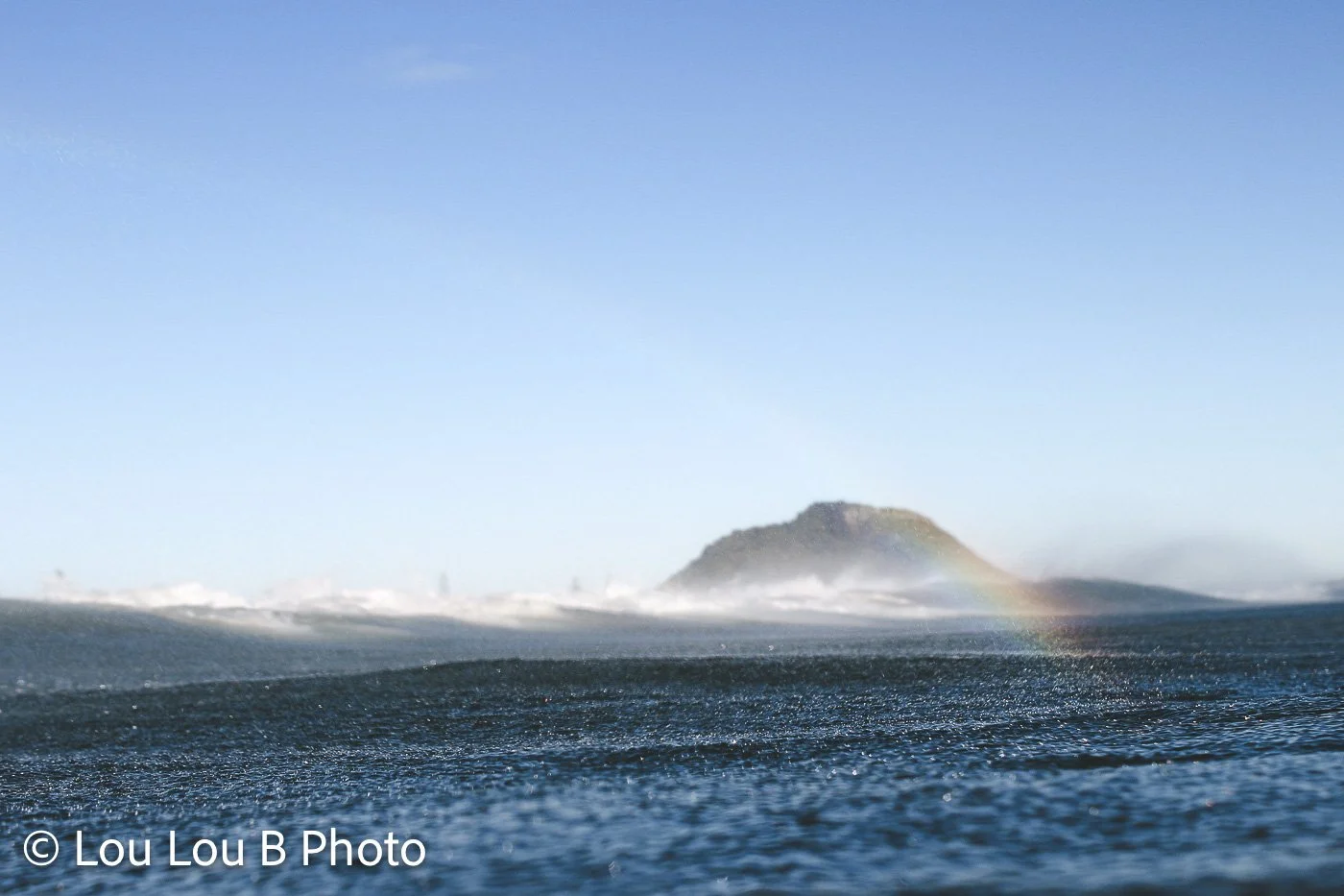 Ocean swell rolling toward a distant island beneath a clear blue sky with sea spray and a faint rainbow.