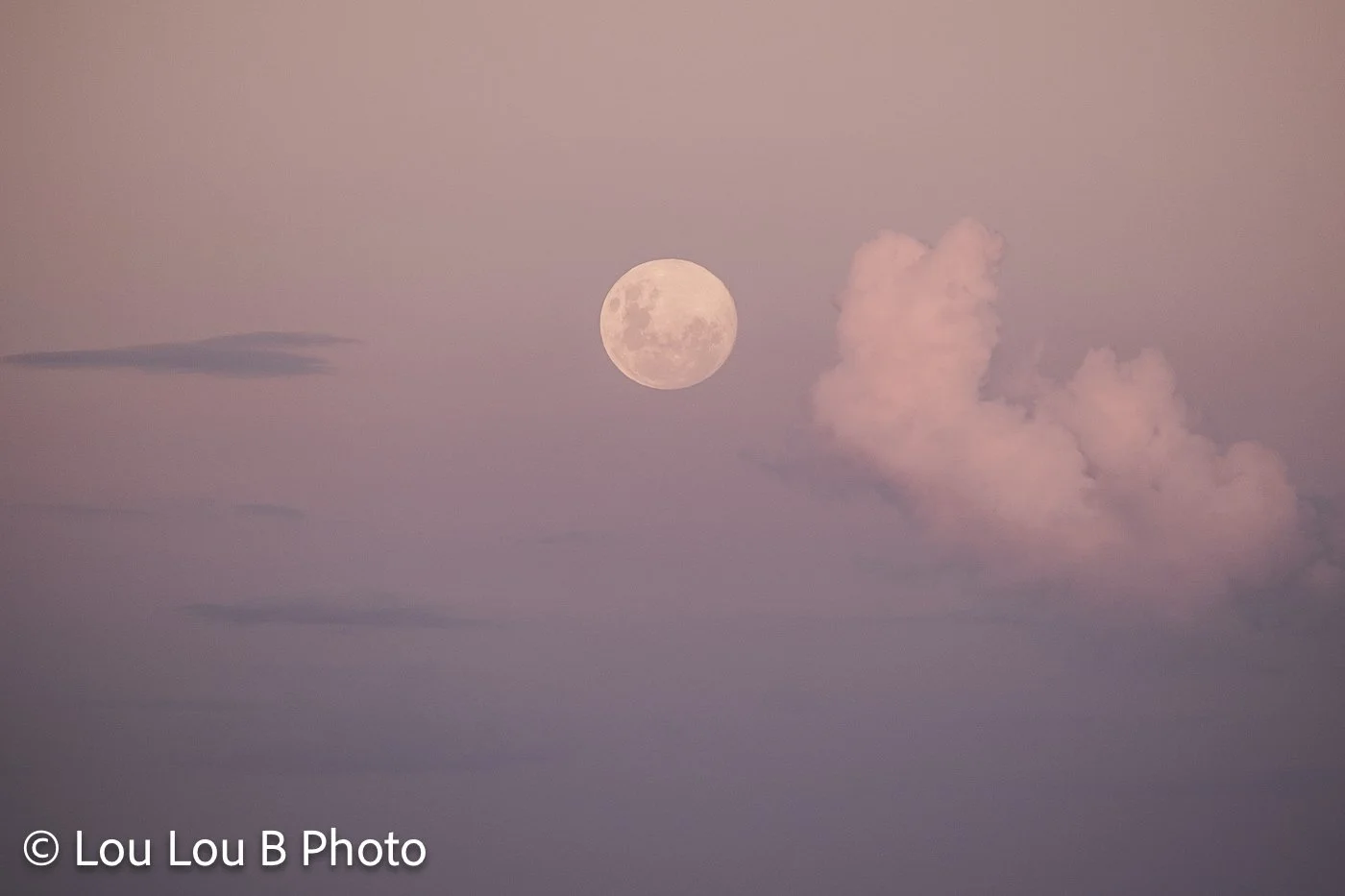 Full moon beside a glowing cloud formation in a soft pink and purple dusk sky.