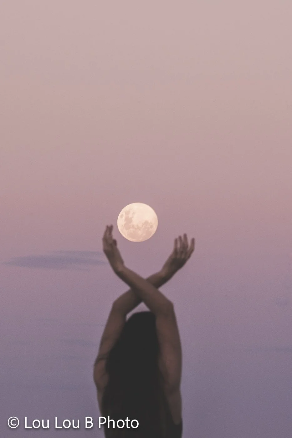 Woman silhouetted against a pastel pink sky, appearing to hold the full moon between her raised hands.