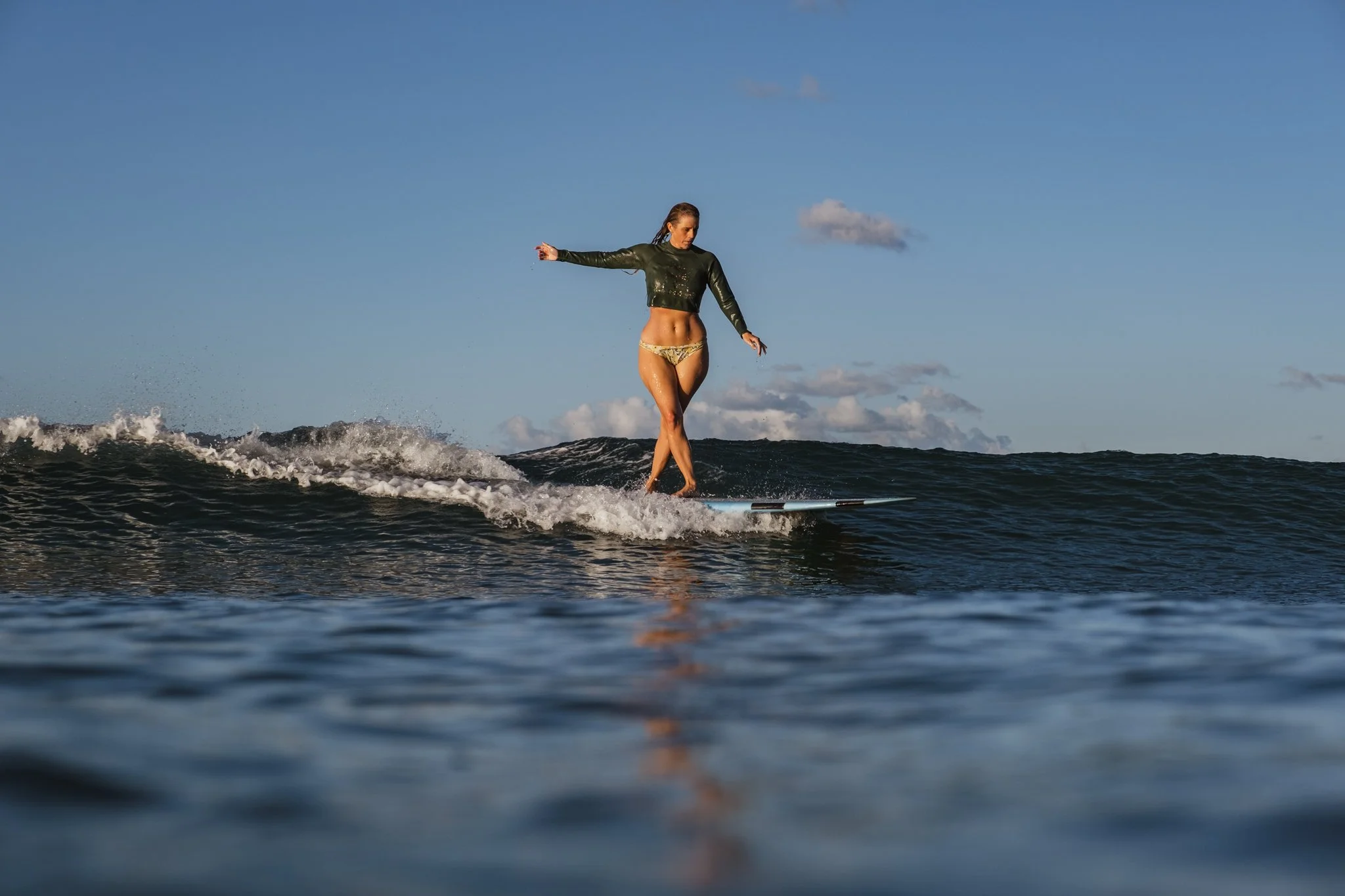 A woman in a dark green Amuse Society cropped rash vest surfs on a longboard in the blue ocean as the warm glow of the evening sun illuminates her. The photo, taken by Lou Lou B, captures crisp water droplets and the woman's fluid motion.