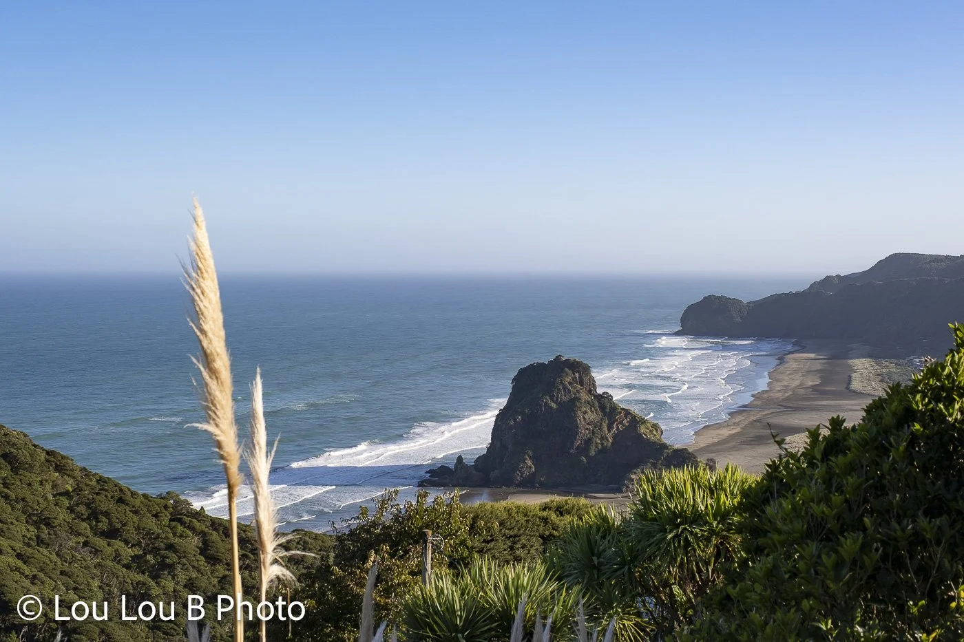 Elevated view of ocean swell rolling toward a rugged black-sand coastline and rocky headland in Piha, New Zealand. 