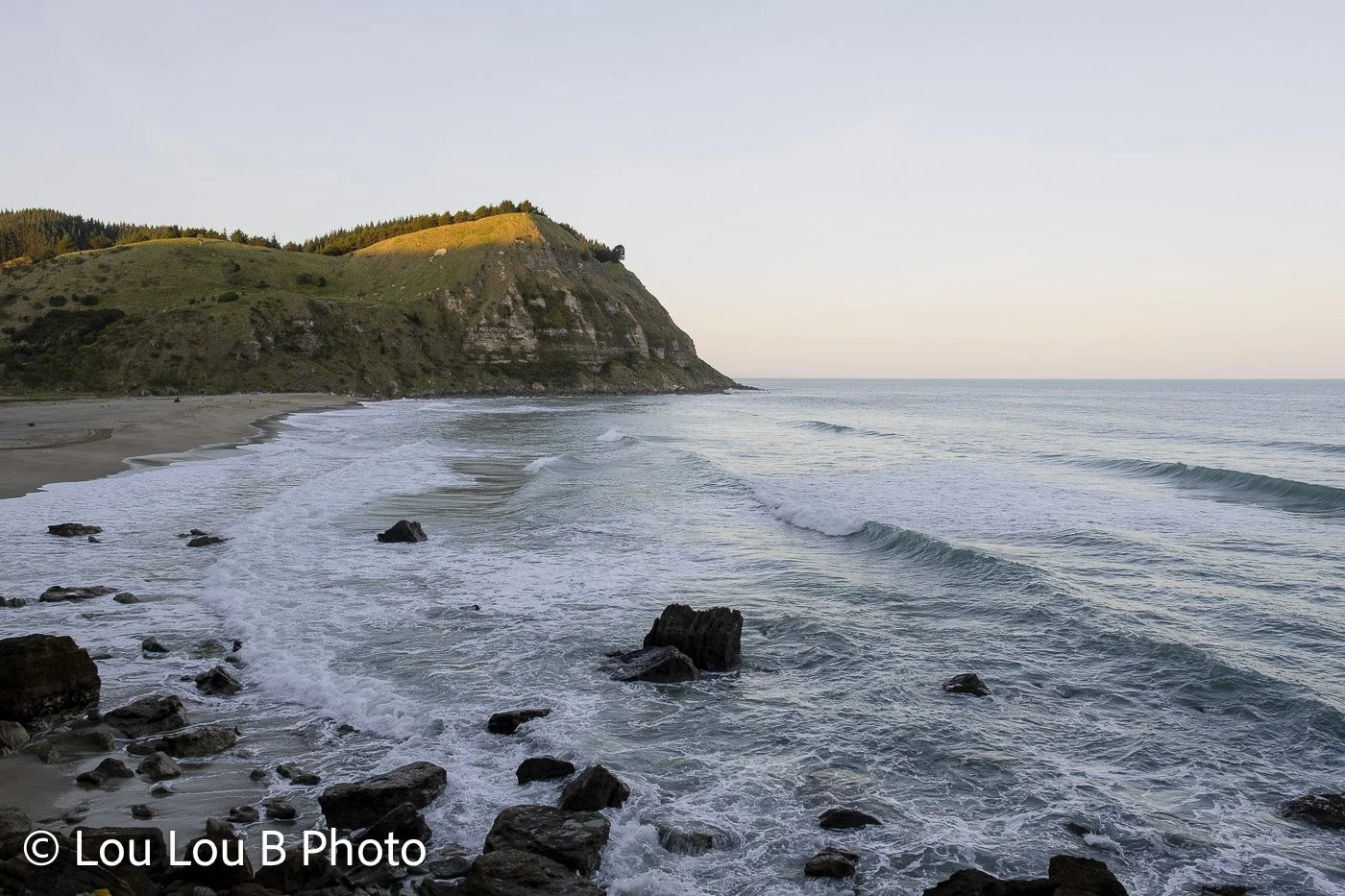 Ocean waves breaking around rocks along a rugged coastal headland in soft evening light.