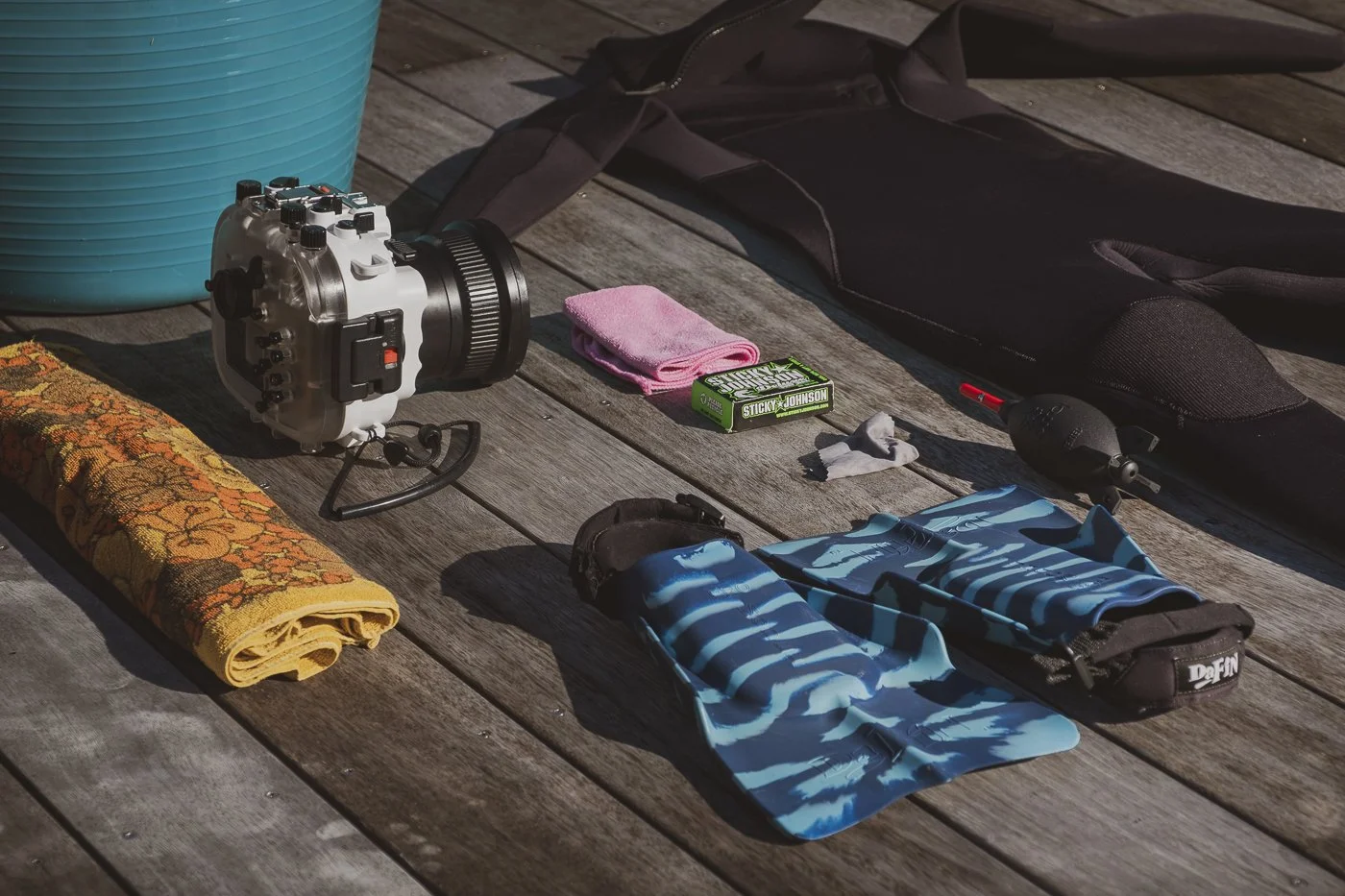 Mirrorless camera in a waterproof housing beside swim fins, wetsuit and surf photography gear on a wooden deck before an ocean session.