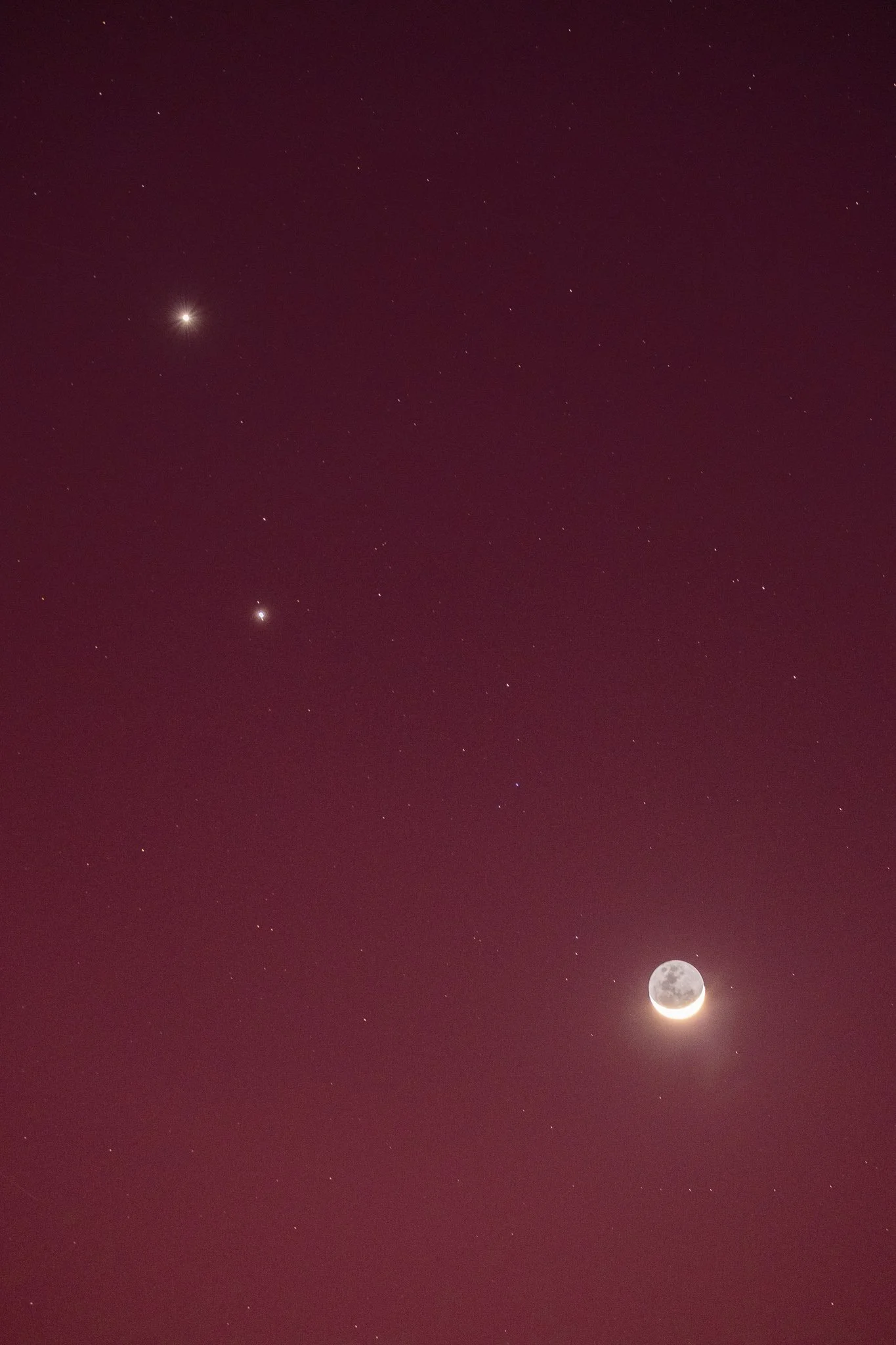 A beautiful pre-blue hour sky frames a stunning composition of the crescent moon in the lower right and bright Venus in the top left. The rich crimson pink hue complements the serene and captivating display of the night sky.
