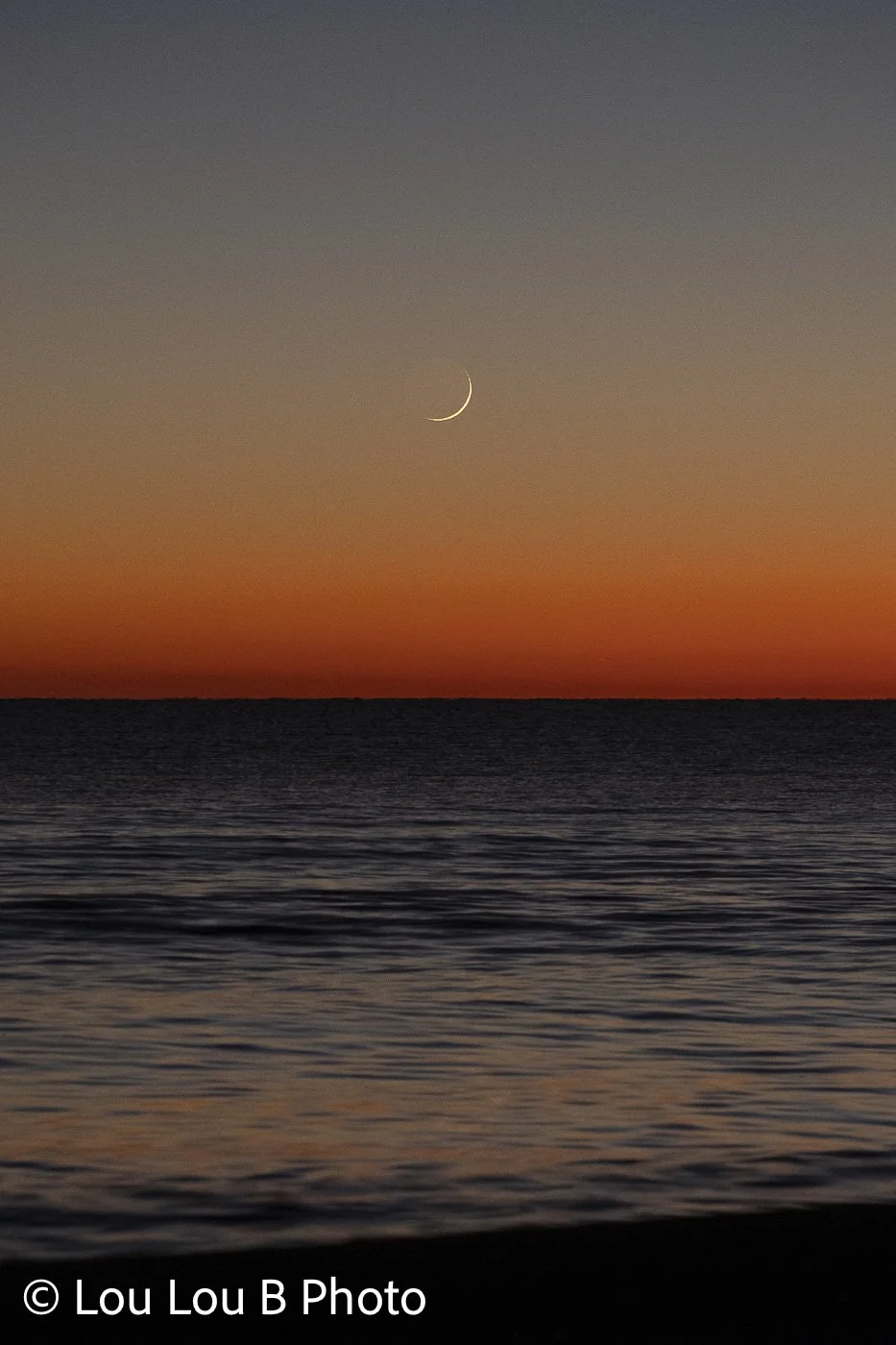 Fine crescent moon above the ocean horizon beneath a soft blue and orange twilight sky.