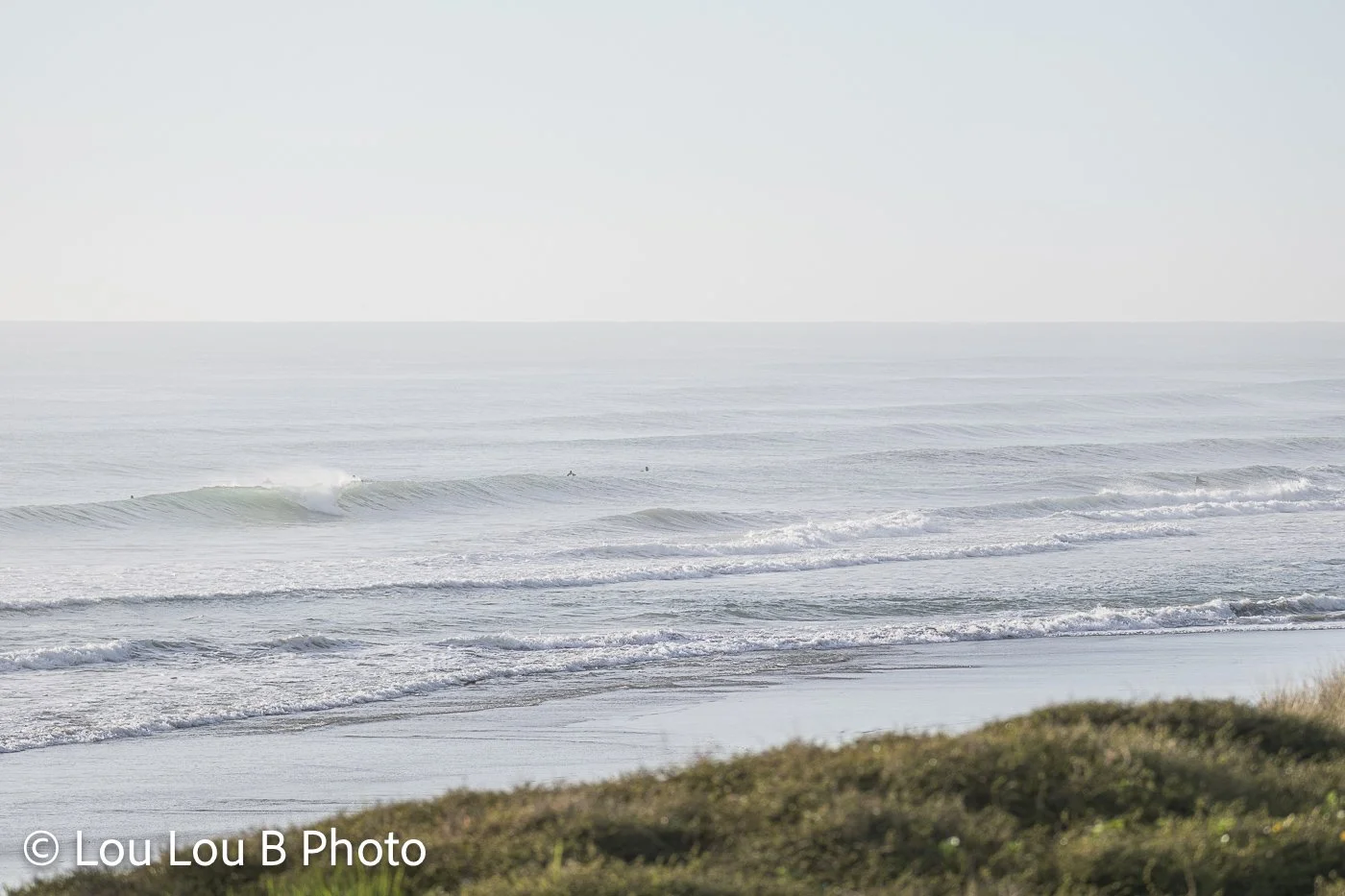 Wide view of soft silver-blue waves rolling toward a quiet coastline beneath a pale sky.