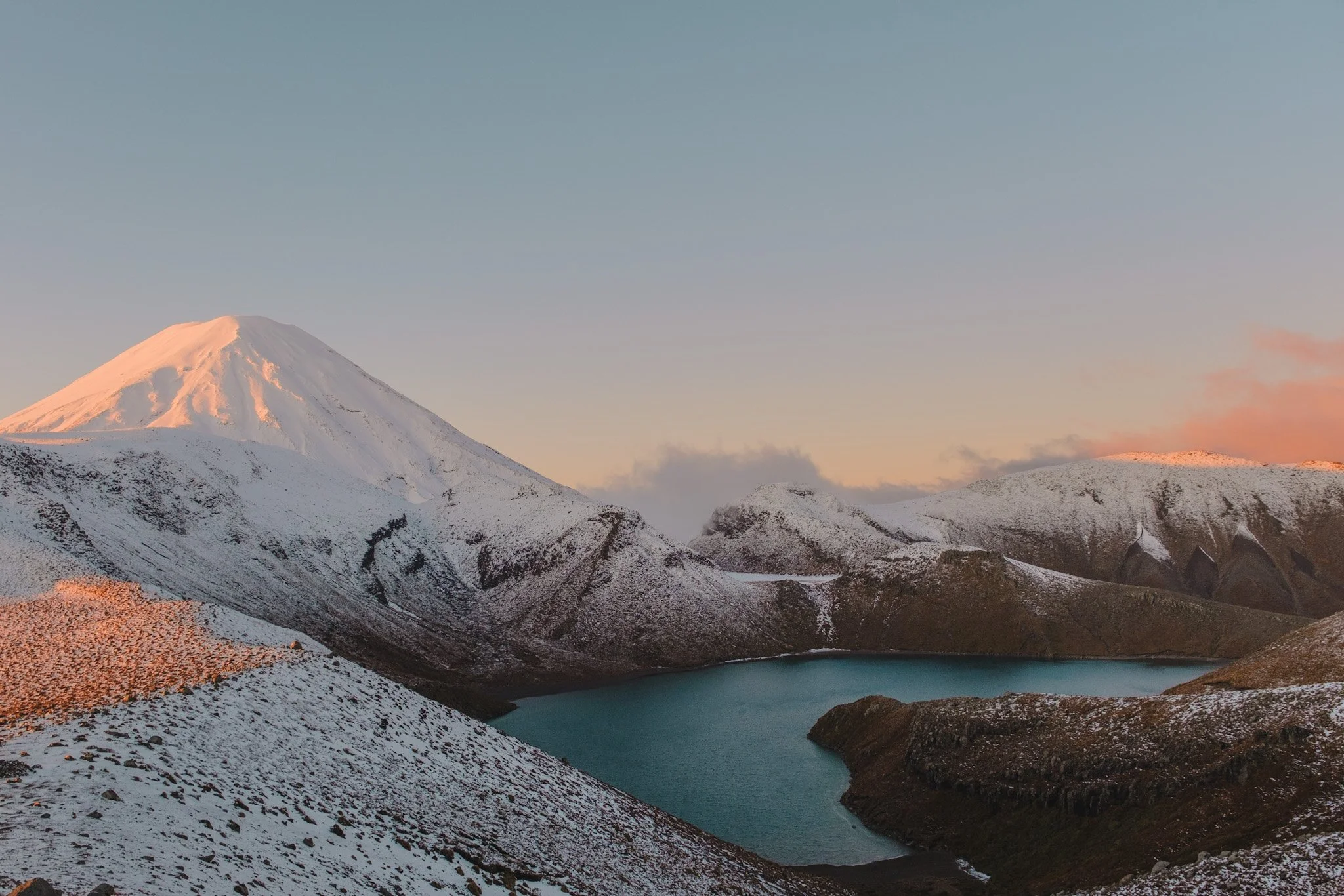 Experience the stunning beauty of New Zealand's Tongariro Crossing with this captivating landscape photograph by Lou Lou B Photo. The golden sun sets behind the snow-capped Mount Ngauruhoe, casting a bright pink light over the Tama Lakes. The rugged 
