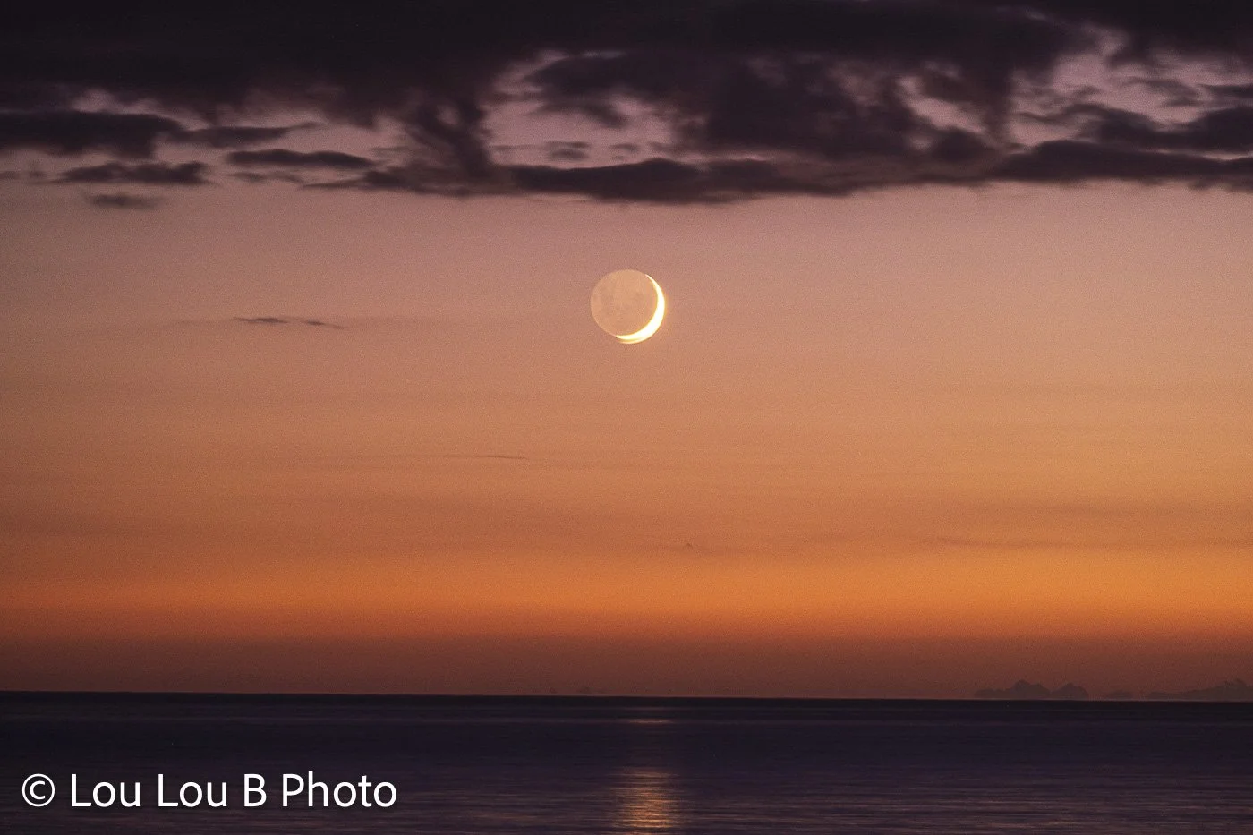 Crescent moon above the ocean beneath dramatic clouds and a glowing peach-orange twilight sky.