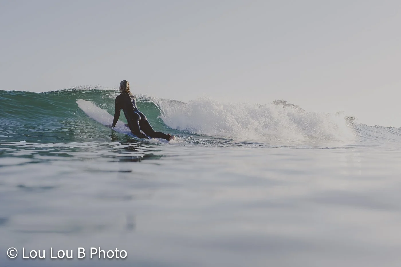 Surfer paddling toward a breaking turquoise wave viewed from water level.