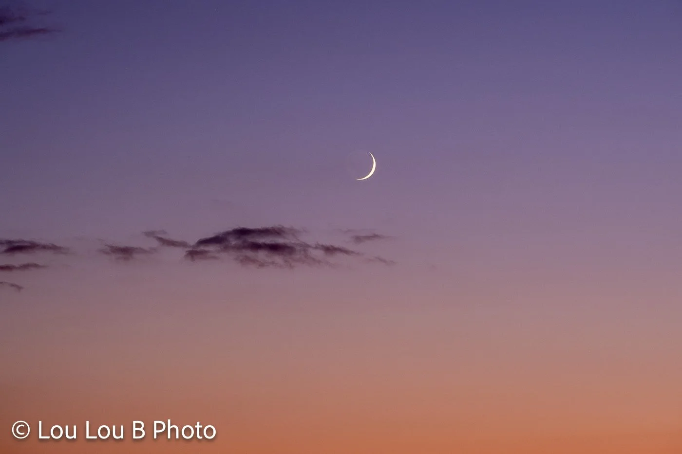 Fine crescent moon above wispy clouds in a pastel purple, pink and orange evening sky.