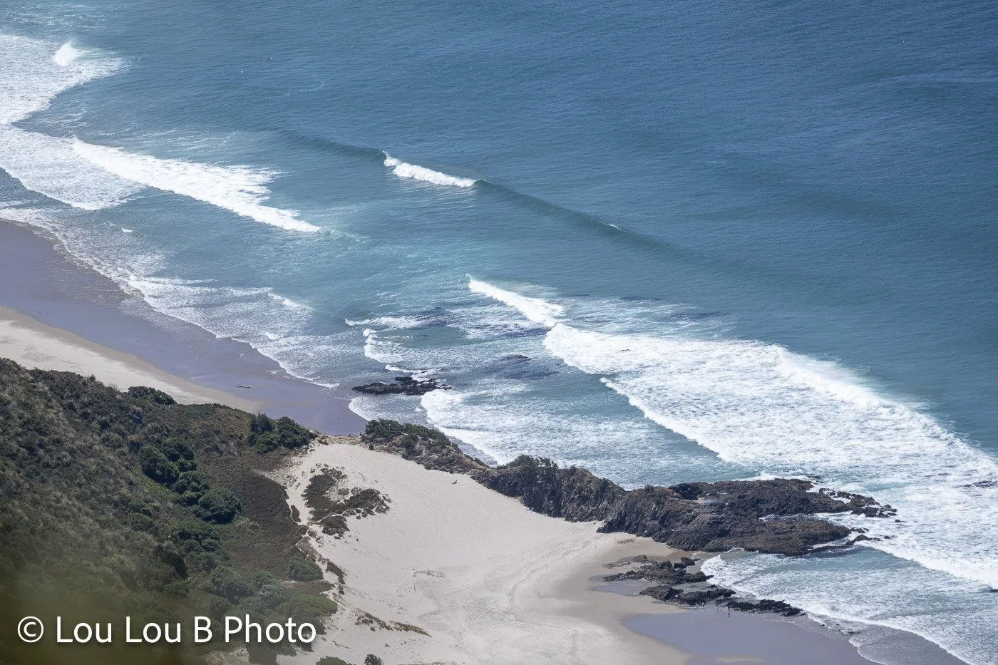Aerial view of powerful ocean swell breaking in parallel lines along a rugged sandy coastline.