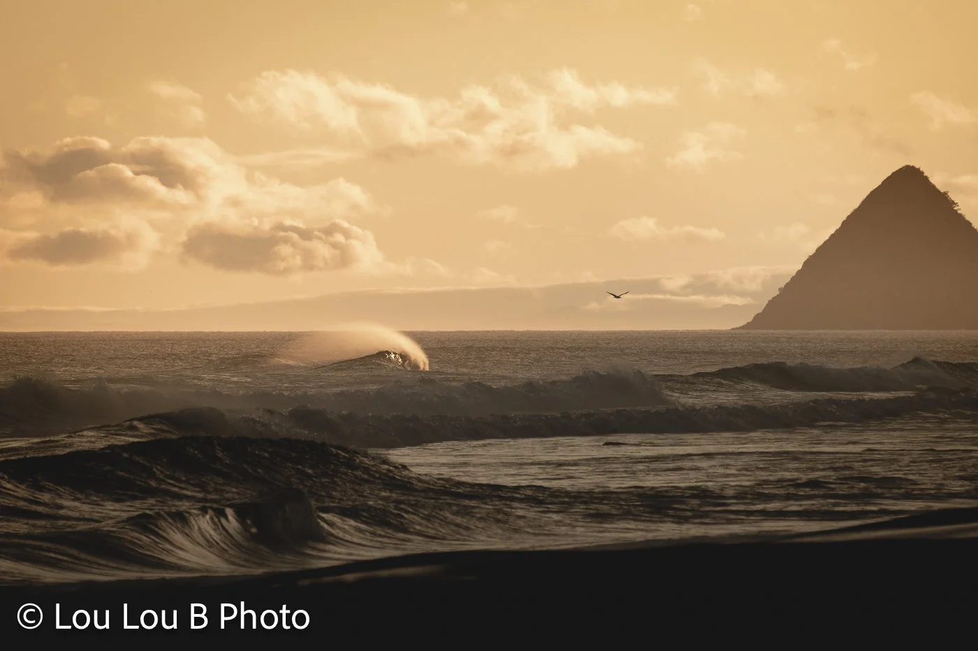 Dramatic ocean swell and offshore spray beneath a golden sky with a steep coastal mountain in the distance.