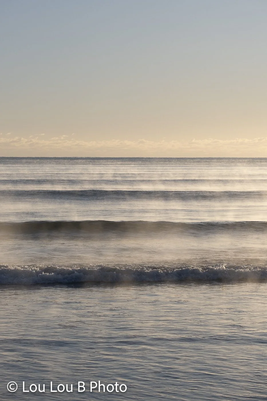 Gentle ocean waves rolling toward shore beneath a pale blue and warm cream sky.
