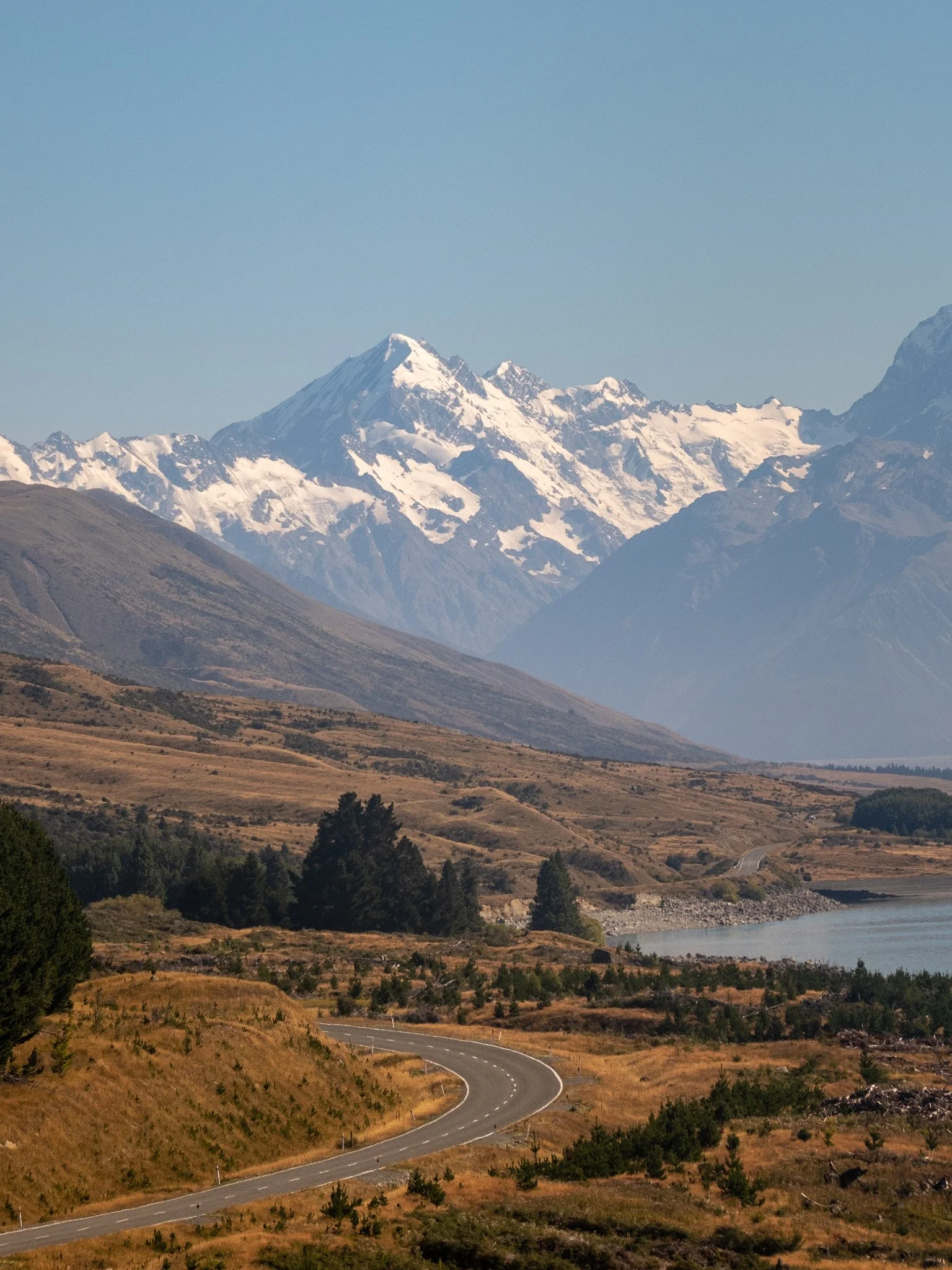 Portrait photo of Mount Cook viewed from State Highway 80, near Lake Pukaki in New Zealand. The winding road leads towards the majestic snow-capped mountain on a sunny summer day, with clear blue sky and alpine landscape in the foreground.