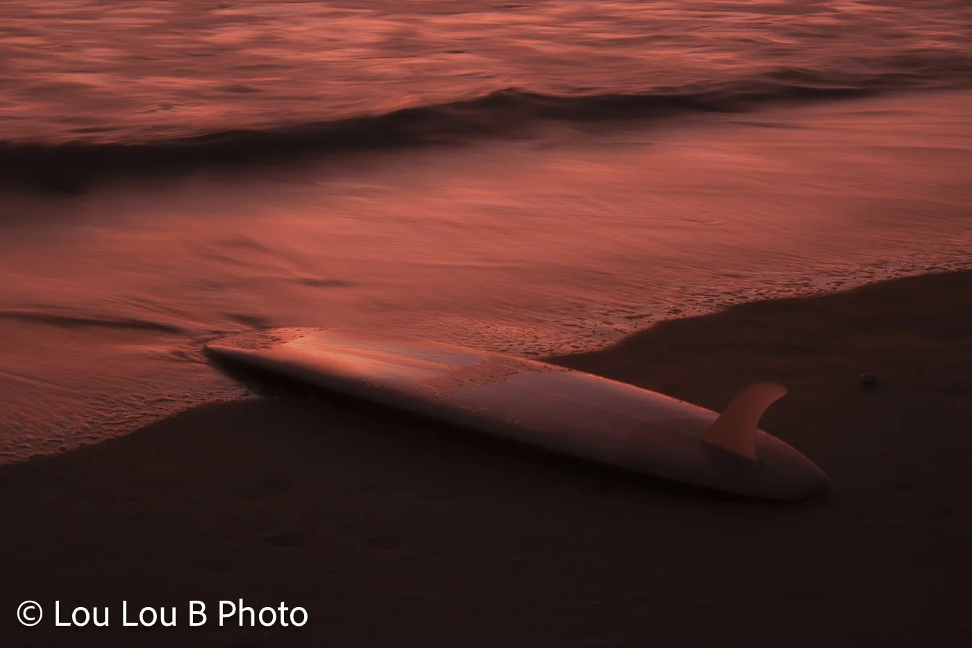 Surfboard resting on a dark beach beside the incoming tide beneath deep red sunset light.