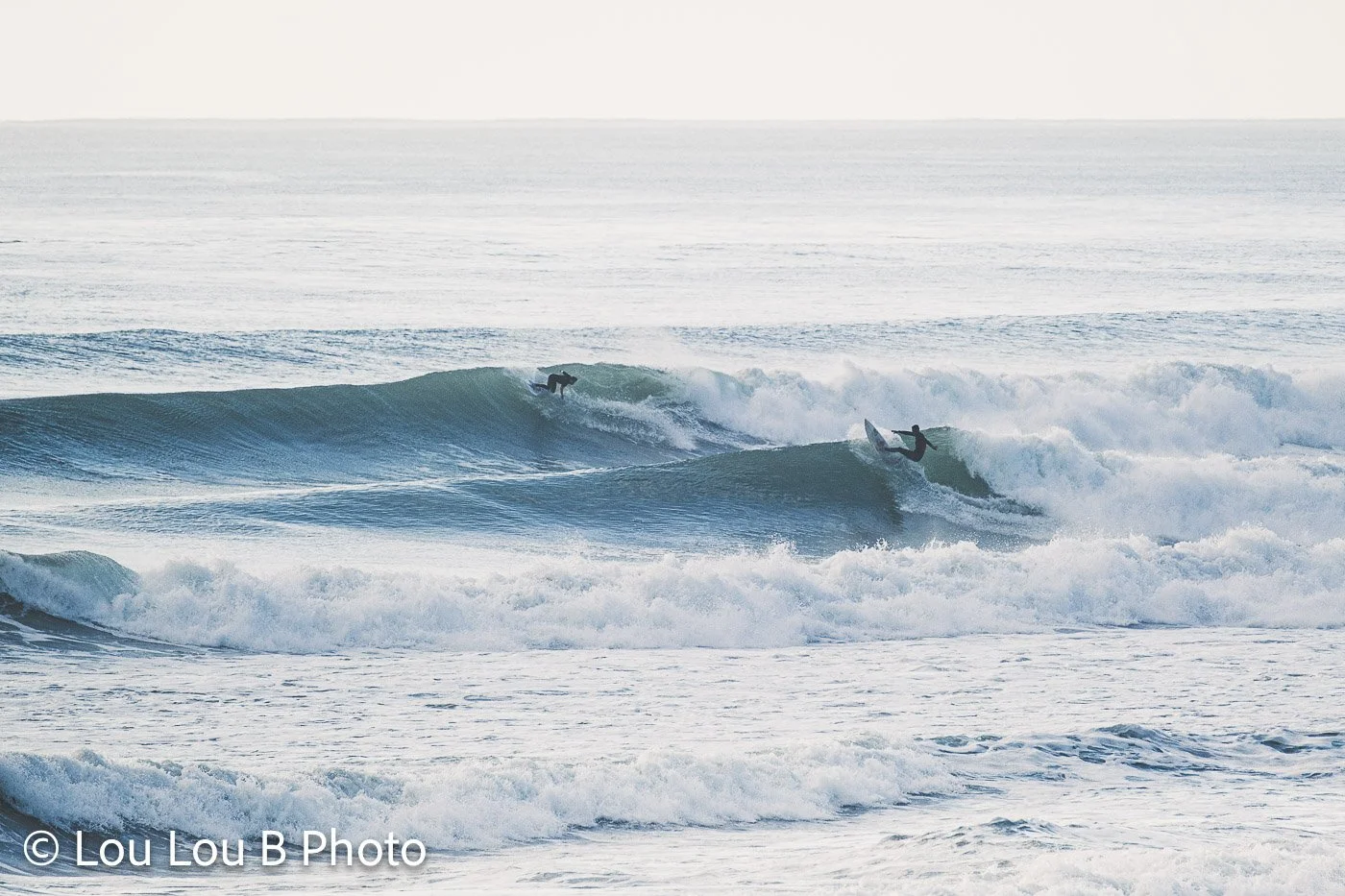 Surfers riding a breaking wave surrounded by whitewater with other surfers waiting beyond.